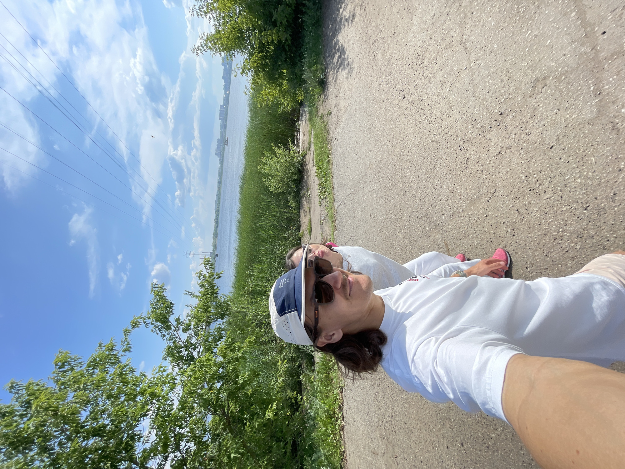 A man and a woman are walking on a paved path next to a body of water in Izhevsk, Russia. The man is in the foreground, wearing a white long-sleeved shirt, a white and blue baseball cap, and sunglasses. The woman is behind him, wearing a white outfit and pink sneakers. The path leads towards a body of water with tall green reeds growing along the edge. In the background, there are trees, power lines, and buildings on the opposite shore. The sky is blue with scattered clouds.