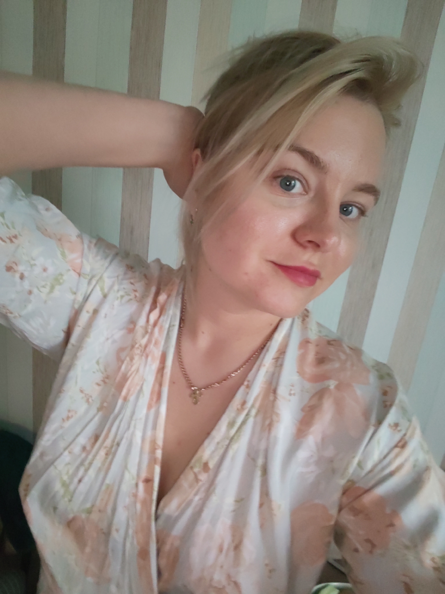 A woman is in a room with wallpaper featuring vertical stripes of white, tan and green. She is fair-skinned with blonde hair and blue eyes, wearing pink lipstick and a gold chain. The woman is wearing a floral print robe with white, tan and green colors. Her left arm is raised with her hand touching the back of her head. The image's location is not provided.