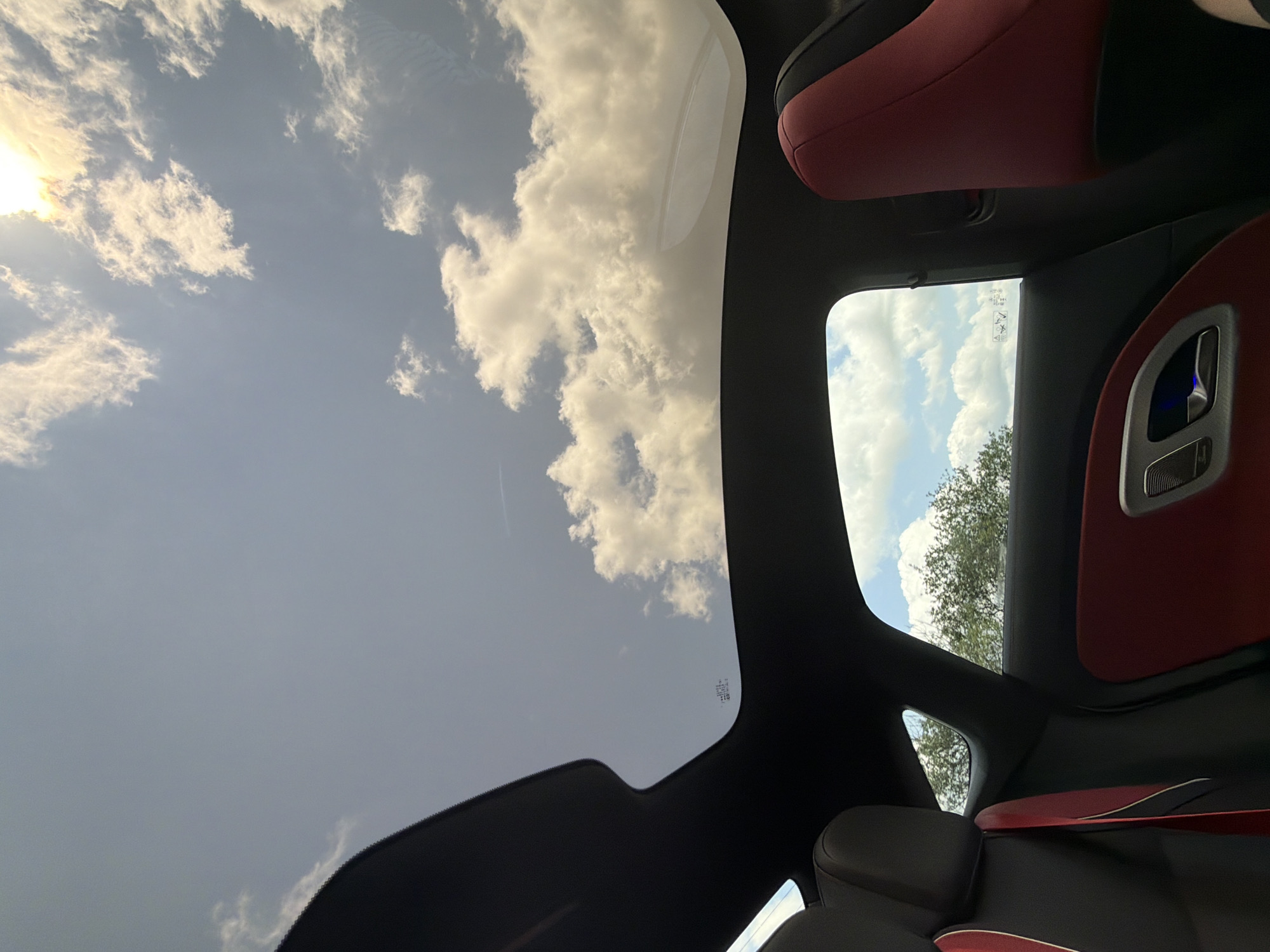 The image presents a view from inside a modern car, looking upwards through a panoramic sunroof and sideways through a window at the sky. The interior features a combination of black and red upholstery, with dark fabric on the headliner and plush red accents on the seats and door panels. A silver door handle with a subtle blue light is visible on the right. A red seatbelt with a white stripe also contributes to the car's interior aesthetic.
Above the car, the sky is partly cloudy with scattered, bright white cumulus clouds against a pale blue background. The sun is visible and bright in the upper left corner of the frame, partially obscured by clouds, indicating daytime. Through the side window on the right, green foliage from a tree is visible, suggesting the car is parked or driving near natural surroundings.
The weather appears fair and bright. No specific activities are depicted other than the observation of the outside world from within the vehicle. There are small, illegible markings or text on the glass of the sunroof, likely manufacturing or safety information. The image does not contain any visual cues to specifically confirm the location as Sadovets, Bulgaria, other than the provided context.