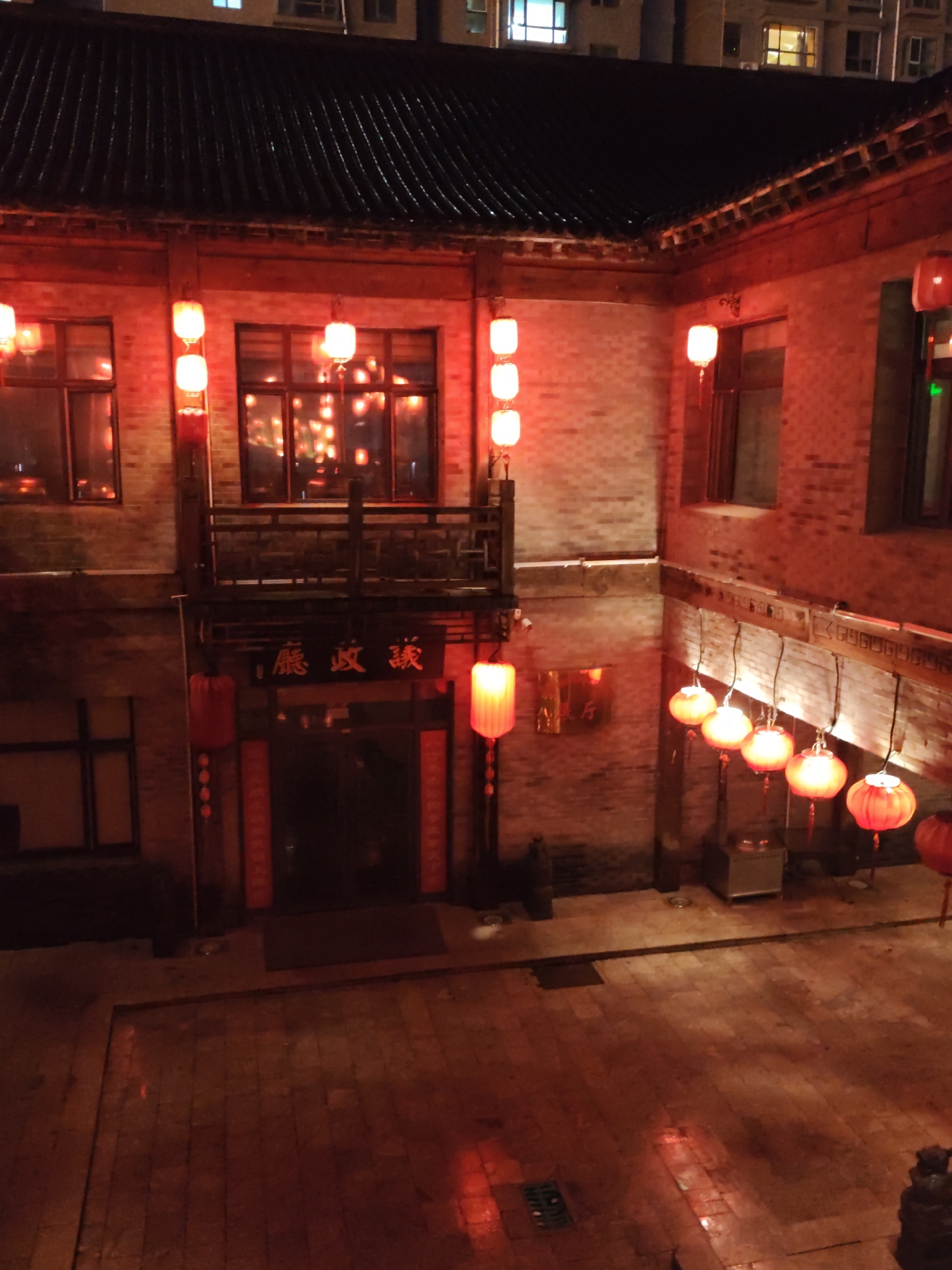 The image depicts a courtyard at night, illuminated by numerous red and yellow lanterns. The architecture suggests a traditional Chinese style, with dark, tiled roofs and brick walls. On the upper floor, there is a balcony overlooking the courtyard, accessible by stairs. Several windows on the upper and lower levels are visible, some reflecting the lantern light. A doorway in the center of the lower level has Chinese characters above it, which translate to "Longzheng Tea House". Various red lanterns hang from the eaves and balcony, as well as on poles in the courtyard. The ground is paved with stone tiles and shows reflections of the lights. In the background, the upper floors of modern buildings with illuminated windows are visible, indicating an urban setting. The overall scene conveys a festive and traditional atmosphere. The location is identified as Lindian, China. There are no people visible in the image.