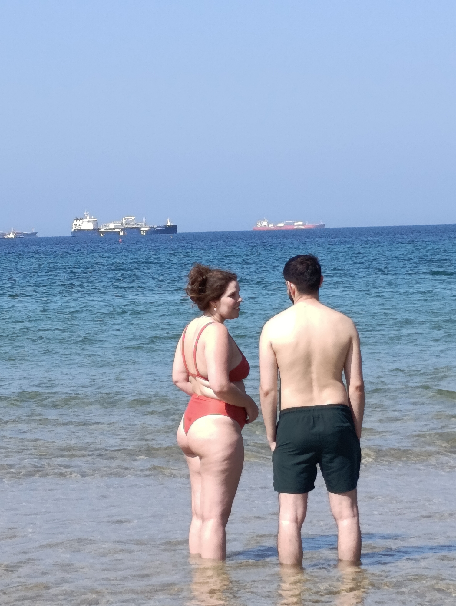 Two people, a woman and a man, stand in the shallow water of a beach, facing away from the camera and towards the sea. The woman, on the left, wears a red bikini. The man, on the right, wears dark green swimming trunks. In the background, several cargo ships are visible on the horizon against a clear blue sky. The scene appears to be in Sines, Portugal, an area known for its port and coastal scenery.