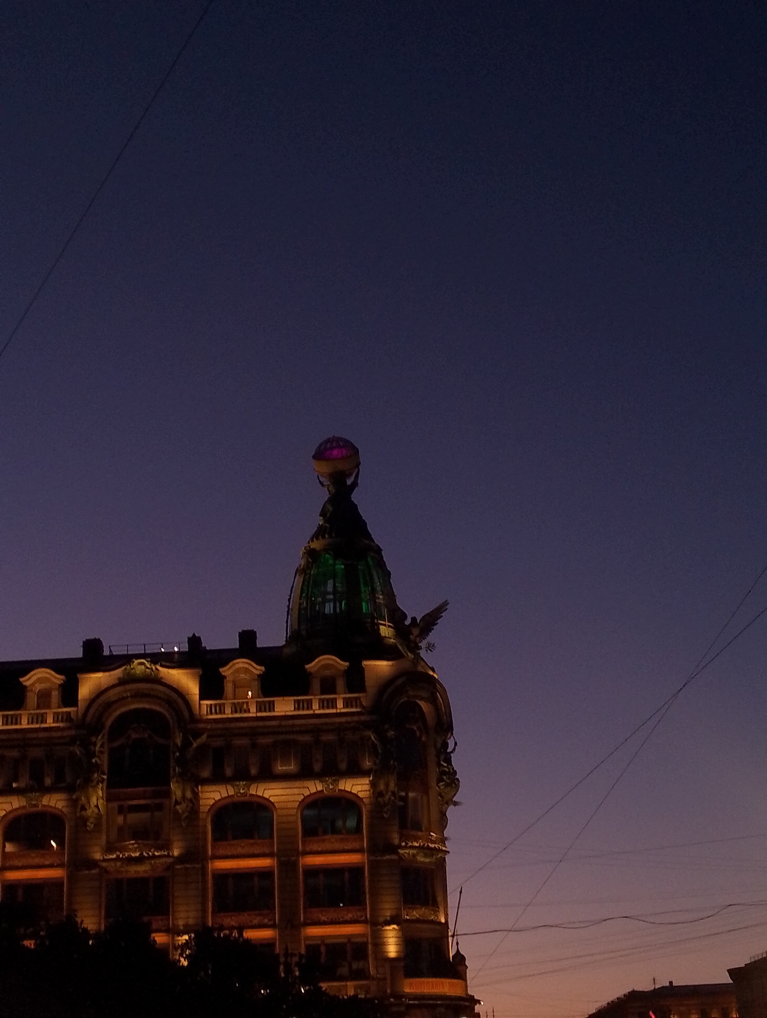 The image shows the Singer House, also known as the Dom Knigi, in Saint Petersburg, Russia, at twilight. The building's ornate facade is illuminated by warm lights, highlighting its arched windows and decorative elements. A prominent glass dome, topped with a sculpture of an eagle holding a globe, crowns the building. The sky is a deep indigo, transitioning to a lighter, hazy pinkish-orange near the horizon. Several utility wires are visible crisscrossing the sky. No people are visible in the scene.