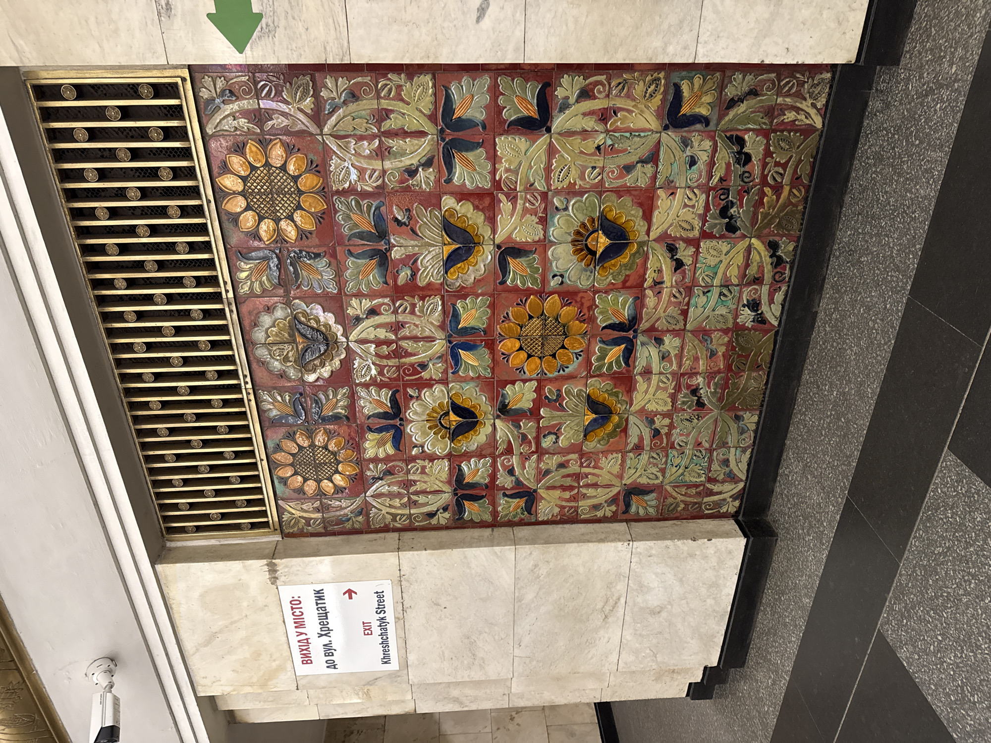 The image shows a wall adorned with decorative tiles featuring floral motifs in red, yellow, blue, and green. A sign with Cyrillic text indicating "EXIT to Khreshchatyk Street" is mounted on a beige tiled wall to the left of the decorative tiles. A security camera is visible on the ceiling above the sign. A green arrow pointing left is visible on the wall to the right of the decorative tiles. The floor is covered in dark grey speckled tiles and black tiles. The scene appears to be in a public indoor space, likely a metro station or underpass, in an urban environment.