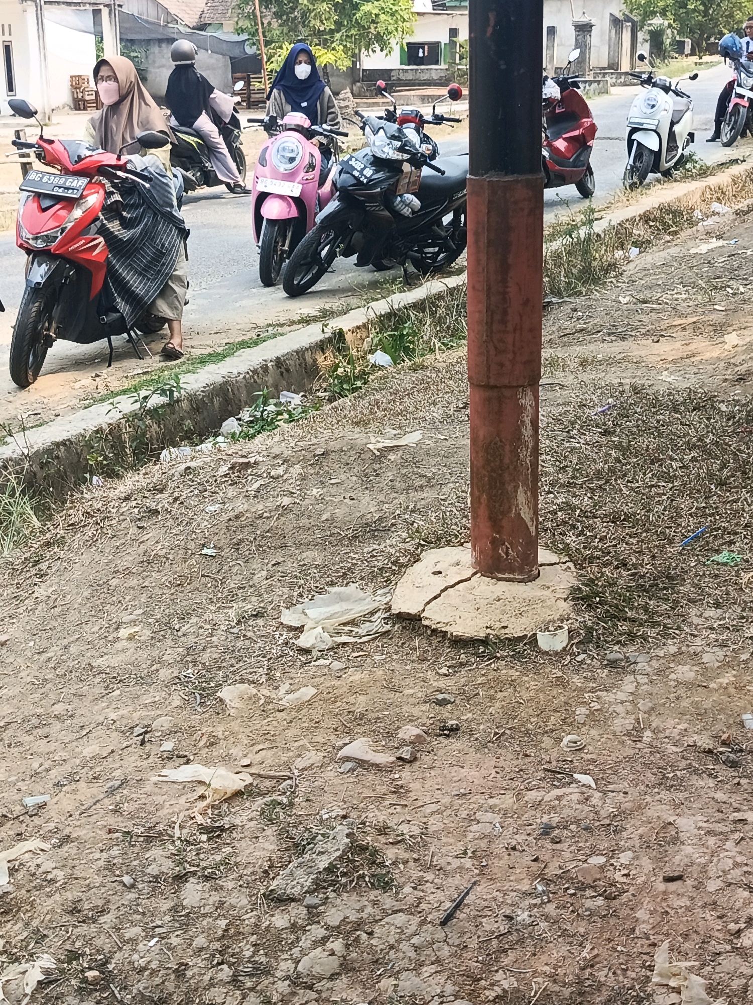 The image depicts a scene on a roadside in Wonosari, Indonesia, featuring several motorcycles and individuals. In the foreground, a red Honda motorcycle is parked, with a person wearing a brown hijab and a striped garment seated on it. To the right, a pink scooter is visible, with a person in a blue hijab standing beside it. Further down the road, other motorcycles, including a white scooter and a dark-colored motorcycle, are parked. The environment appears to be dusty and unkempt, with debris scattered on the ground and a drainage ditch running alongside the road. The overall impression is of a typical street scene in a provincial Indonesian town.