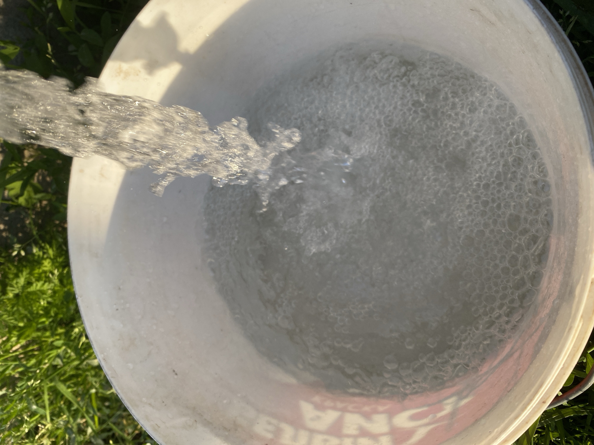 A white bucket is positioned on green grass, with water pouring into it from above. The water creates many bubbles and ripples as it enters the bucket, suggesting it is being filled. The bucket has some pink lettering on its side, but it is partially obscured by the water. The scene is outdoors, with sunlight illuminating the water and grass.