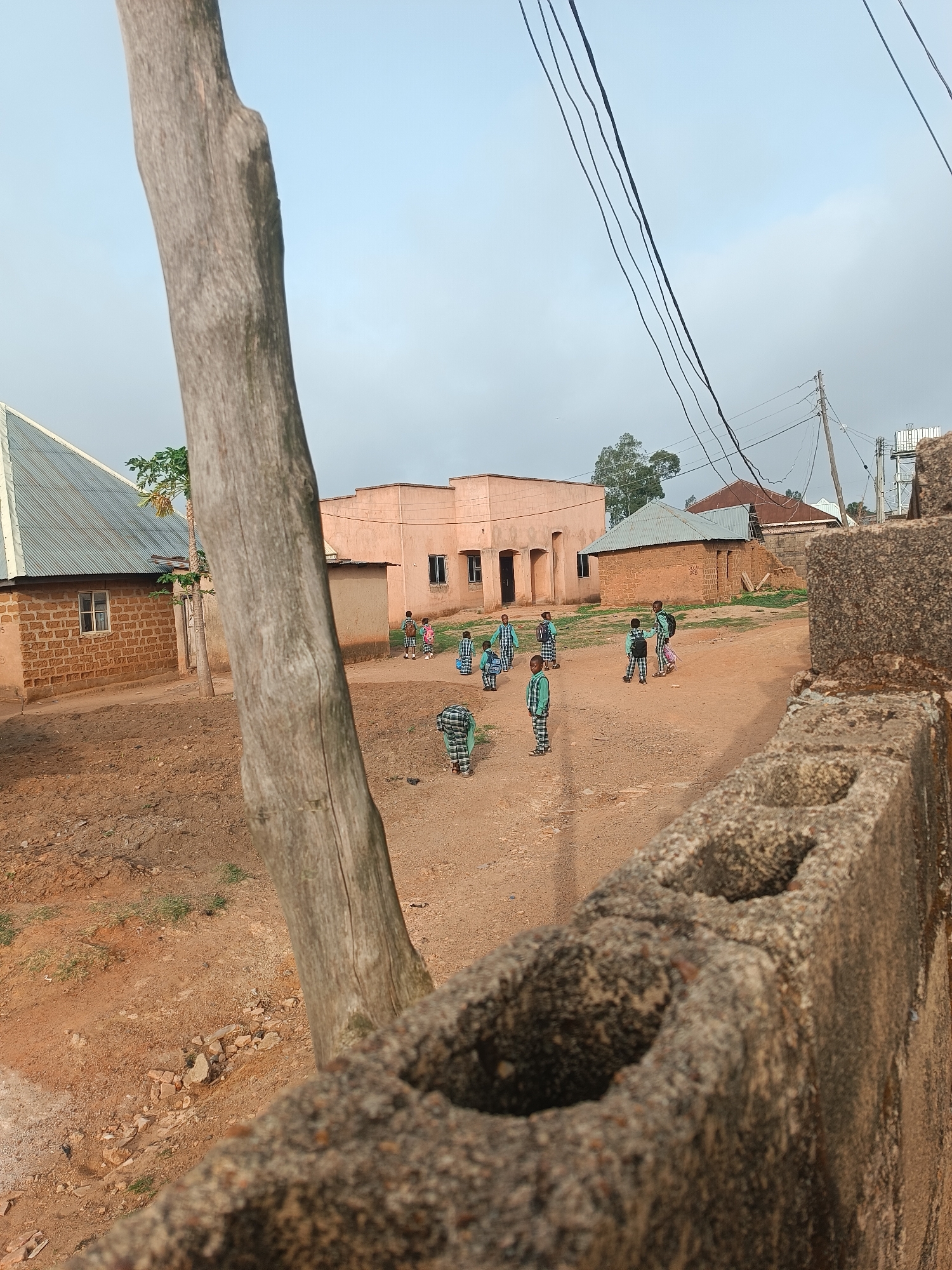 The image depicts an outdoor scene in Bukuru, Nigeria. A group of children, numbering around nine, are scattered in a dusty, unpaved area between several buildings. The children are wearing what appear to be school uniforms. The buildings are primarily one-story structures constructed of brick or block, with corrugated metal roofing. A bare tree trunk is prominently positioned in the foreground on the left, while a concrete block wall is on the foreground right. Power lines stretch across the sky above the buildings. The sky appears overcast or hazy.