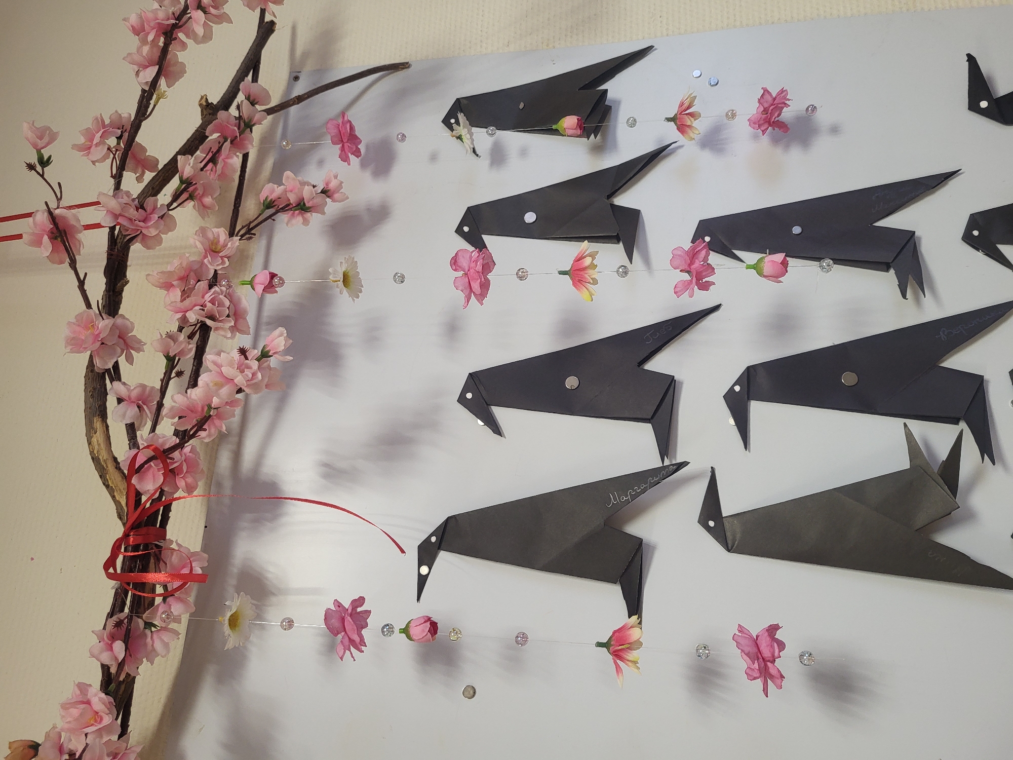 The image presents a white surface on which several black origami birds are mounted, each seemingly affixed with a small, silver-colored fastener. Most of the birds have writing on them in what appears to be a Cyrillic script. Strung horizontally across the birds, at approximately beak-level, is a thin line adorned with occasional small, transparent beads and artificial flowers in pink, white, and yellow. To the left side of the frame is a branch covered with artificial pink cherry blossoms and a loosely tied red ribbon.
The location, identified as Chaikovsky, Russia, is based on the Cyrillic script on the birds which suggests a Russian-speaking environment.