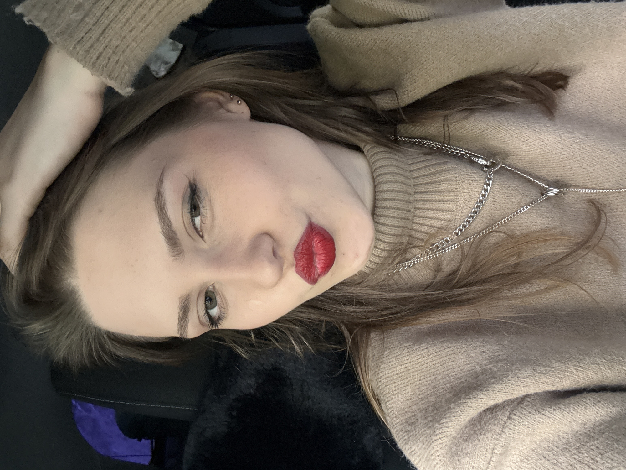 A young woman with fair skin and light brown hair is lying down, facing the camera. Her head is tilted slightly to her left, and she has one hand resting on her head. She is wearing a light beige turtleneck sweater and a silver chain necklace. Her lips are painted a bright red. The background is blurry but appears to include a black surface with a hint of purple. The location is not discernible from the image.