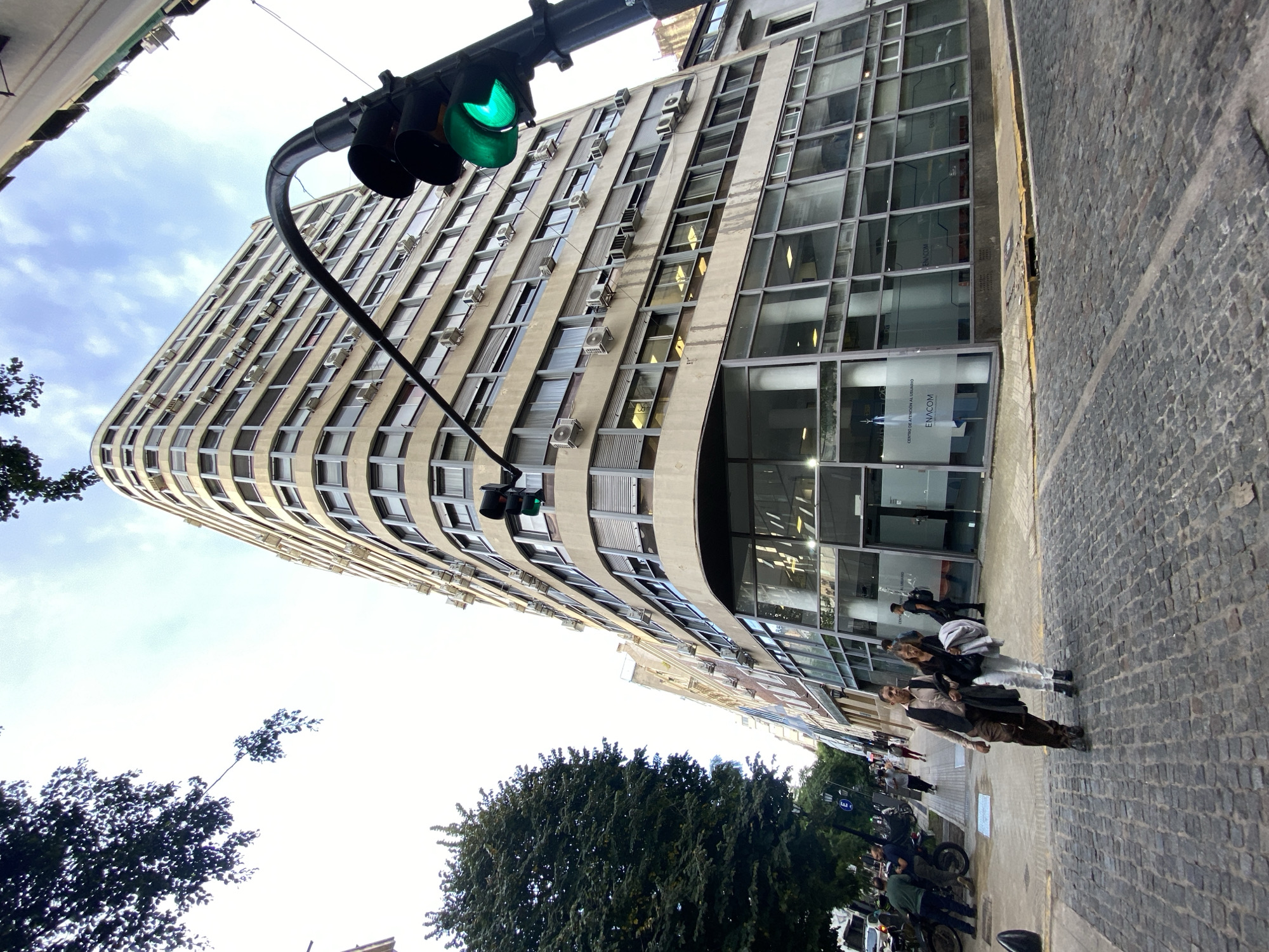 Here's a detailed description of the image:
**Setting:**
The image depicts a street scene in Buenos Aires, Argentina. The street is paved with cobblestones and lined with trees. A tall, curved building with multiple windows dominates the background.
**Objects:**
- A traffic light is visible, displaying a green signal.
- The building has numerous windows, many of which feature small air conditioning units.
- The building's entrance has large glass windows and doors.
- Motorcycles and cars are parked along the curb.
**People:**
- Several people are walking on the sidewalk, mostly near the entrance to the building.
- The people are dressed in casual attire.
**Environment:**
- The sky is partly cloudy.
- The lighting suggests it is daytime.
**Building Location:**
The building is the ENICOM building.