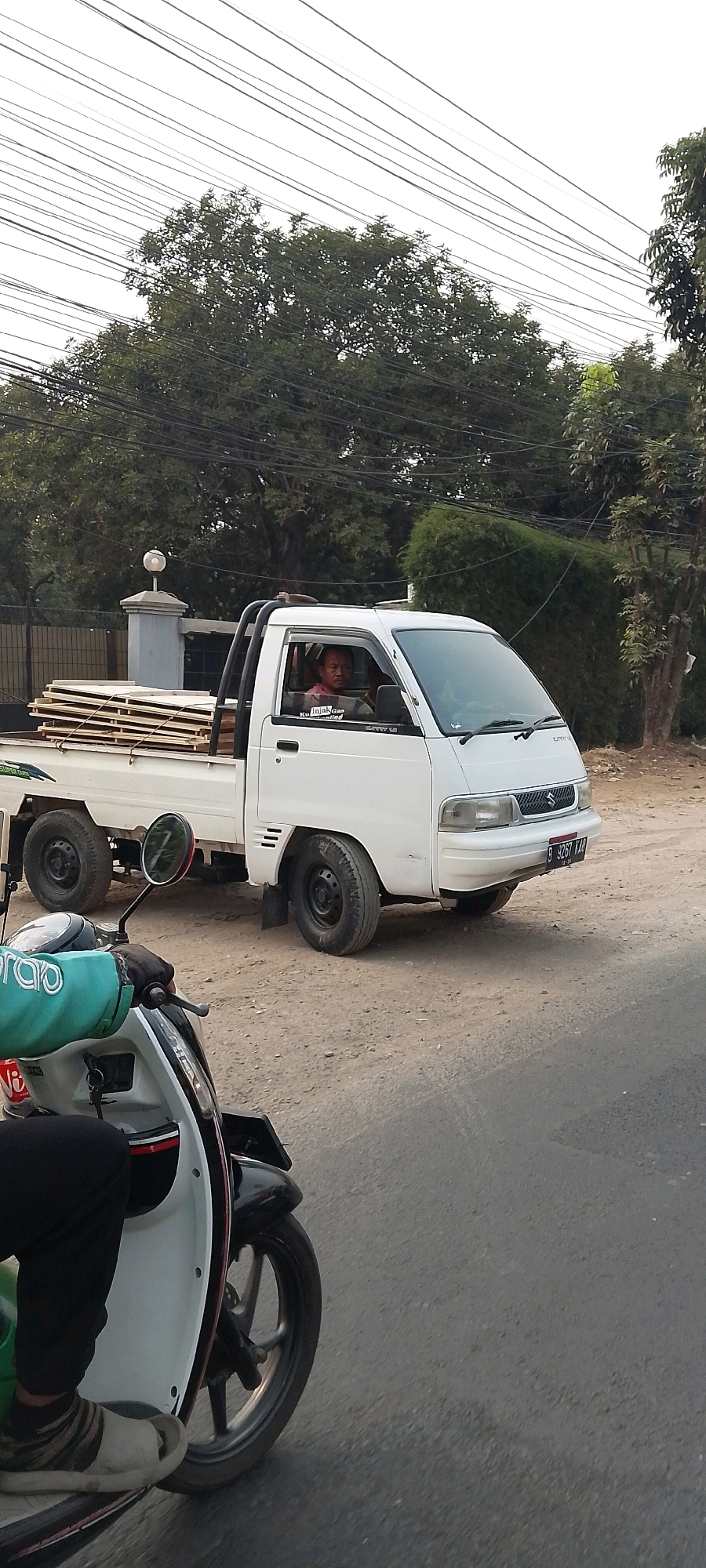 An outdoor, medium-shot, eye-level, slightly angled perspective shows a white Suzuki Carry utility truck parked on a dirt road, with a person riding a scooter in the foreground. The truck's bed is laden with a stack of flat, light-colored rectangular objects, possibly wood planks or panels. A man with dark hair and a light-colored shirt is visible in the driver's seat of the truck, looking forward. The truck has a metal roll bar. In the foreground, a person is riding a white scooter, partially obscuring the truck. The rider's left arm, clad in a green jacket with white lettering that reads "GRAB", is extended, holding the scooter's handlebar. The rider's legs are clad in black pants, and they are wearing striped socks and light-colored sandals. The environment appears to be a roadside in Cileungsi, Indonesia, indicated by a license plate on the truck that reads "B 3267 KAJ" and a glimpse of a residential fence and trees. Overhead, numerous dark electrical wires crisscross against a bright sky. The ground is a dusty, light brown dirt.