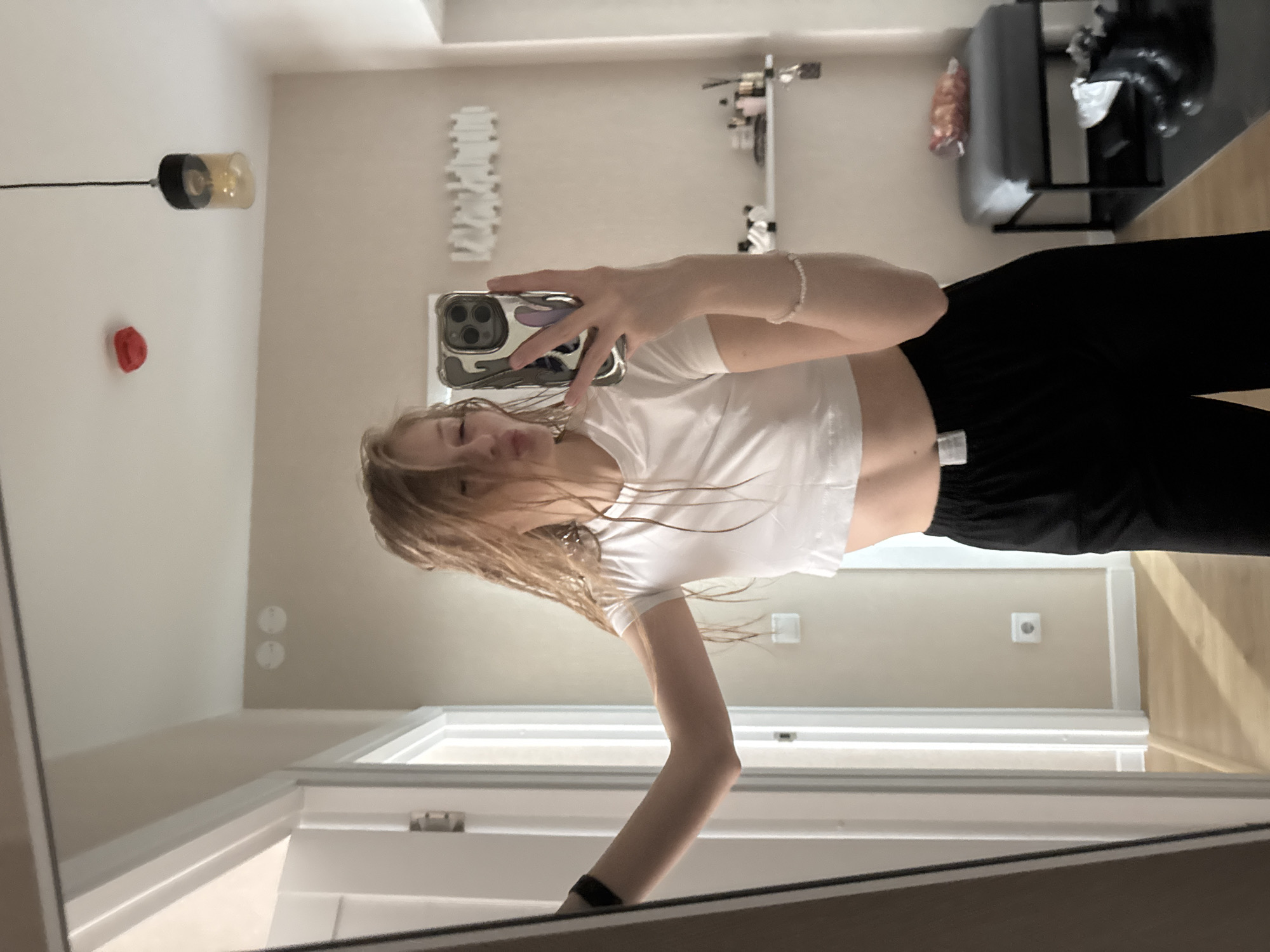 A young woman is taking a selfie in front of a mirror. She is wearing a white cropped t-shirt and black pants. She is holding a smartphone in her right hand. The background shows a room with a light-colored wall, a pendant lamp, and some wall decorations. In the distance, there is furniture including what appears to be a couch or chair. The photo was taken in Perm, Russia.