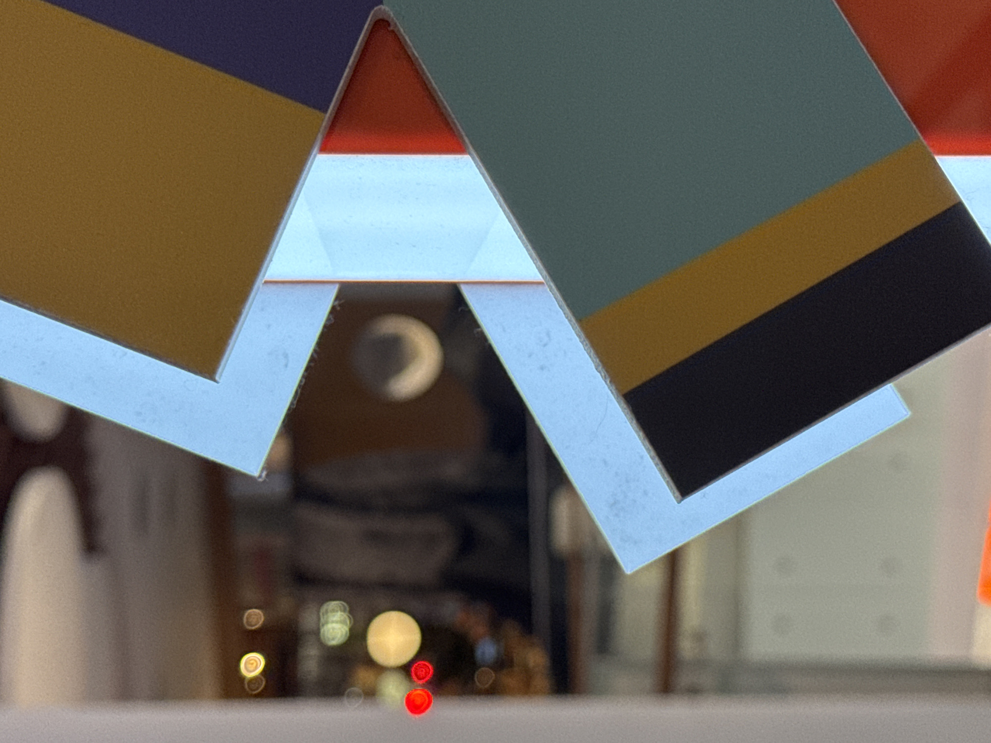 A close-up shot shows overlapping geometric shapes with blurred lights and shadows in the background. The geometric shapes appear to be some kind of display. The shapes have multiple colors, including blue, black, yellow, red and white. In the background, there is a series of out-of-focus lights and reflections, suggesting an urban setting at night. The location is unknown, as city and area information are unknown.