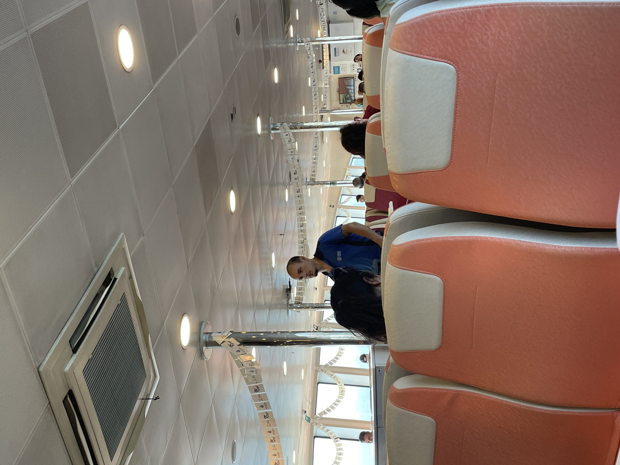 The image shows the interior of a ferry boat, possibly in Üsküdar, Turkey. The scene is viewed from the perspective of a passenger seated in the front rows of orange and cream upholstered seats. In the background, several passengers are visible, including a man wearing a blue shirt standing near a metal pillar decorated with small flags. The ceiling is white and has a grid pattern, with recessed lights and an air conditioning unit. Windows line the side of the ferry, offering a view of the outside. The overall impression is that of a typical public transportation vessel.