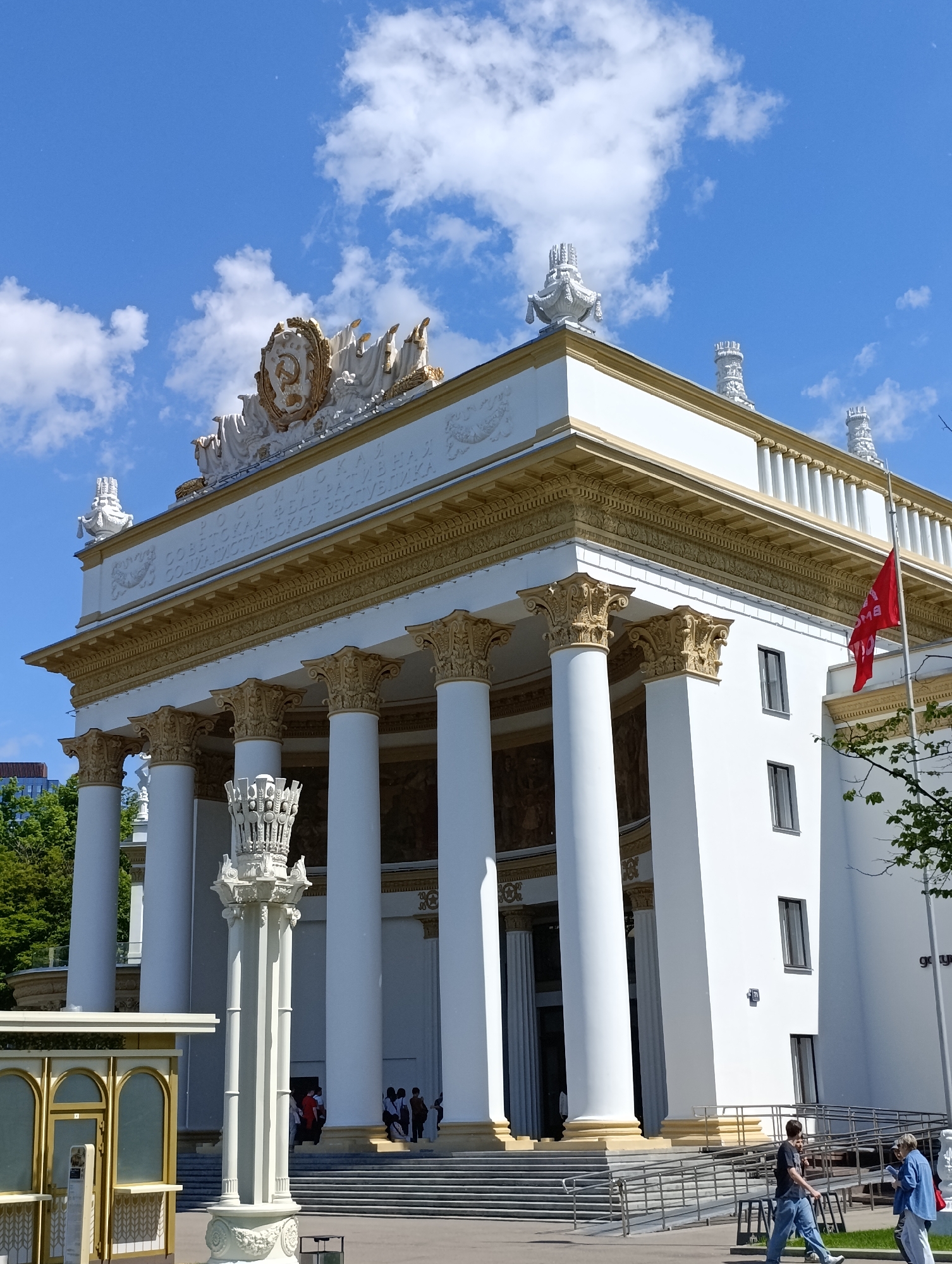 This image showcases a grand, neoclassical-style building under a bright blue sky with scattered white clouds. The building is predominantly white with opulent golden accents, including ornate Corinthian capitals on its tall columns and a richly decorated frieze.
On the frieze, Cyrillic text is visible, partially reading "РОССИЙСКАЯ ФЕДЕРАЦИЯ" (Russian Federation) and "СОВЕТСКАЯ СОЦИАЛИСТИЧЕСКАЯ РЕСПУБЛИКА" (Soviet Socialist Republic), reflecting its historical Soviet-era origin and current context. Crowning the roofline is a prominent golden emblem featuring the hammer and sickle, flanked by various decorative white finials and embellishments. A red flag, with some illegible white Cyrillic text, flies from a flagpole on the right side of the building.
In the foreground, a tall, intricately designed white decorative column or lamp post stands, featuring a crown-like top. To its left, a small, golden-framed kiosk-like structure is present. Several people are visible, some ascending the wide staircase towards the building's entrance, while others walk on the ground level, including individuals near a wheelchair ramp. The scene is bathed in bright daylight, indicating a sunny day. Green trees are visible on the left and right, providing natural accents to the architectural setting.
This building, characterized by its imposing columns and ornate details, is typical of Stalinist architecture, frequently found in Moscow, Russia, and likely represents one of the pavilions at the VDNKh (Exhibition of Achievements of National Economy).