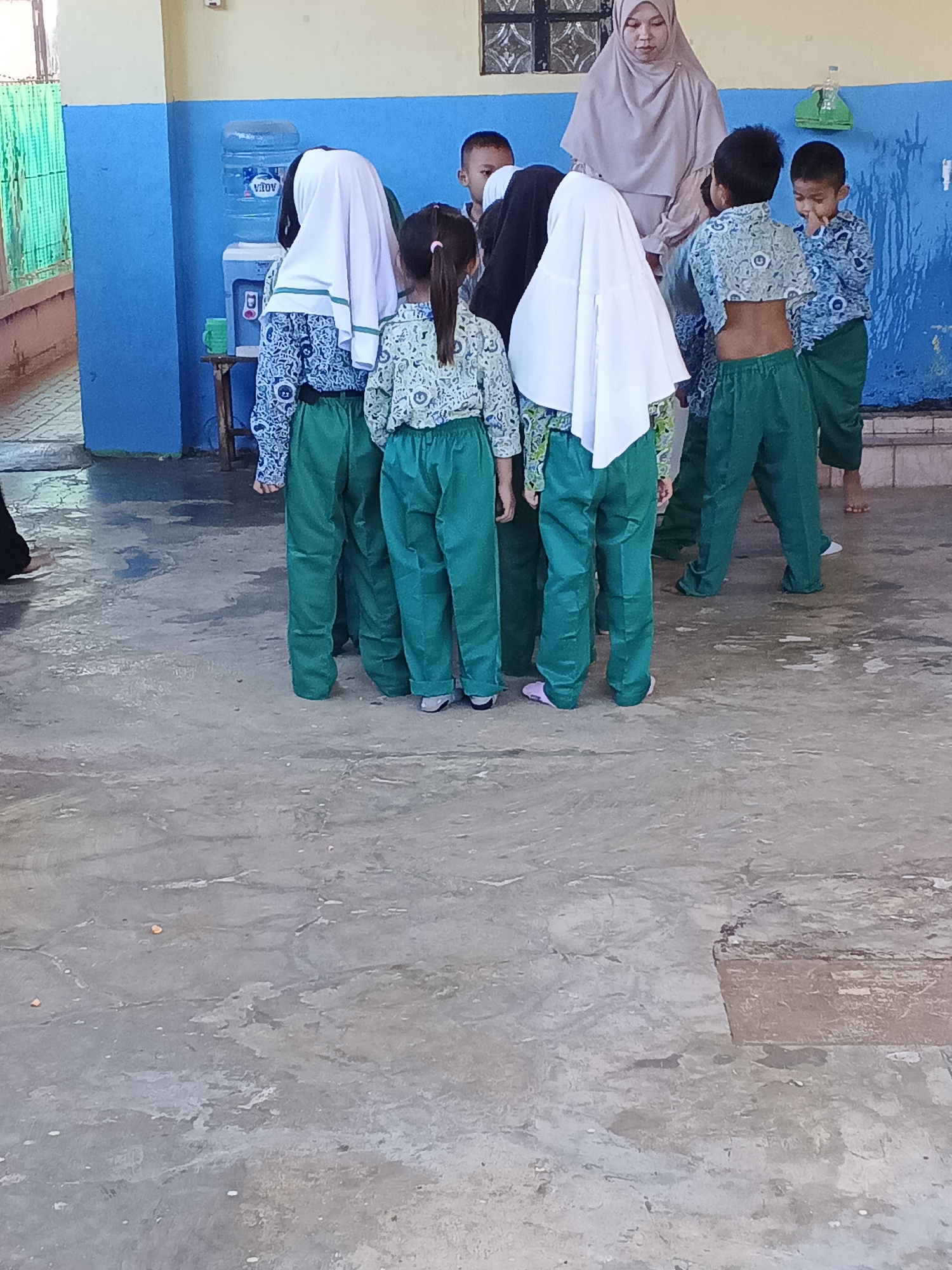 In Warungkiara, Indonesia, a group of schoolchildren in green uniforms stand around a water dispenser. The children, some wearing hijabs, are facing away from the camera. A woman in a beige hijab stands behind them. The scene takes place in what appears to be an outdoor area with a concrete floor and a blue-painted wall.
