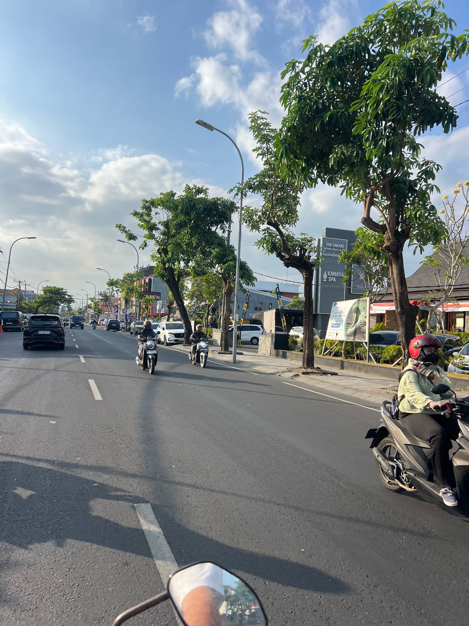 In Kuta, Indonesia, a street scene unfolds under a partly cloudy blue sky. The asphalt road, marked with white lines, stretches into the distance, lined with trees and buildings. Several vehicles are visible: a black SUV, a white car, and multiple motorcycles. Two individuals are riding motorcycles in the foreground. The person on the right, wearing a light green jacket, black pants, a red helmet, and a face mask, rides a dark-colored scooter. The person on the left, also on a scooter, is partially obscured by the vehicle in front. Traffic lights and street lamps line the road, and signs for businesses like "The Sakaye Luxury Villas & Spa" and "Kanaya Spa" are present on buildings. Lush green trees, some with bare branches, are interspersed along the street, and a billboard with an image of a person is displayed on the right side. The overall impression is a bustling street in a tropical urban environment.