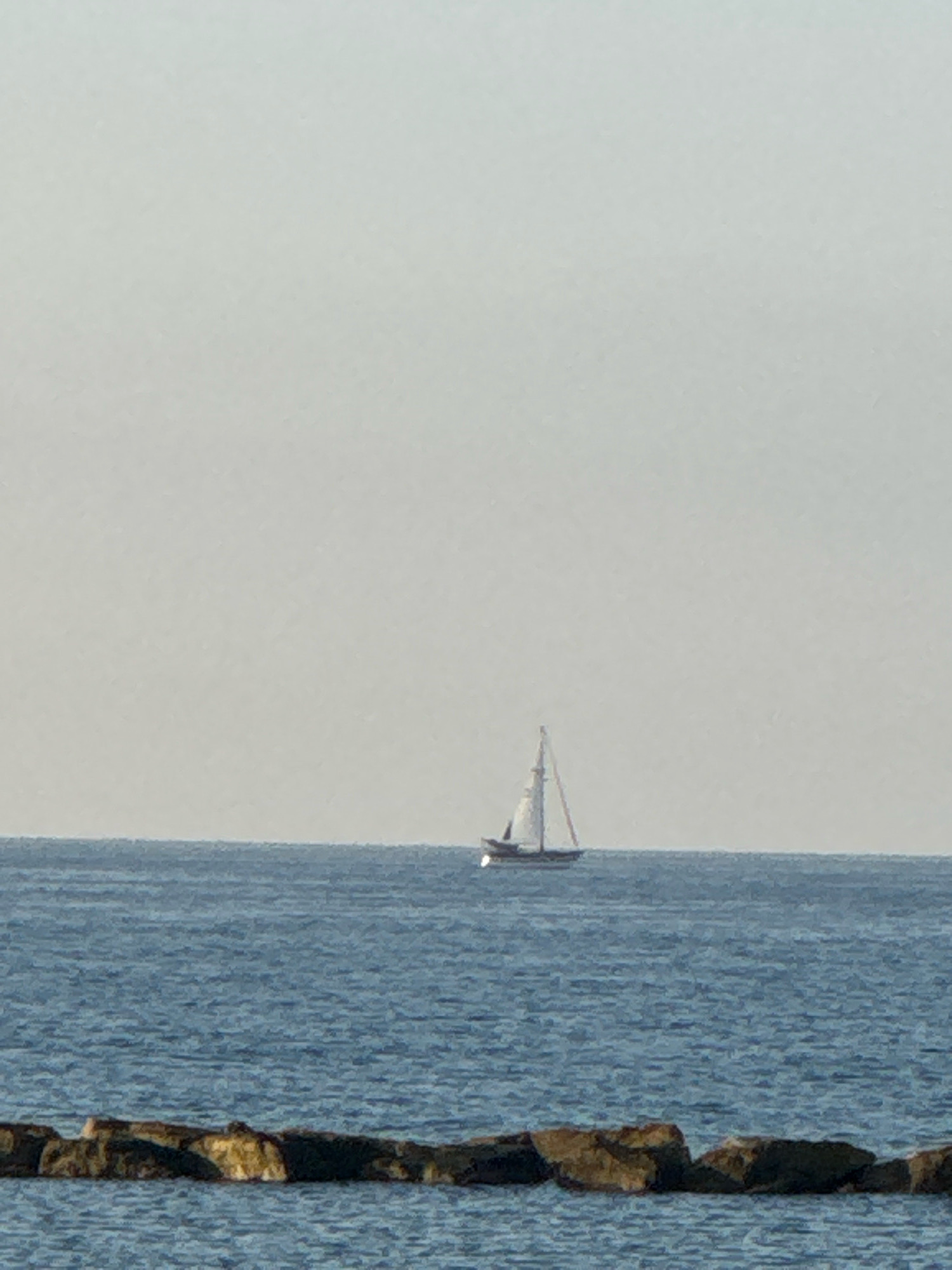 A sailboat with white sails is sailing on a calm, blue sea. The sky is a pale, hazy gray. In the foreground, a line of large, dark rocks is visible partially submerged in the water. The scene is serene and evokes a sense of peacefulness. The location is Agios Tychona Community, Cyprus.