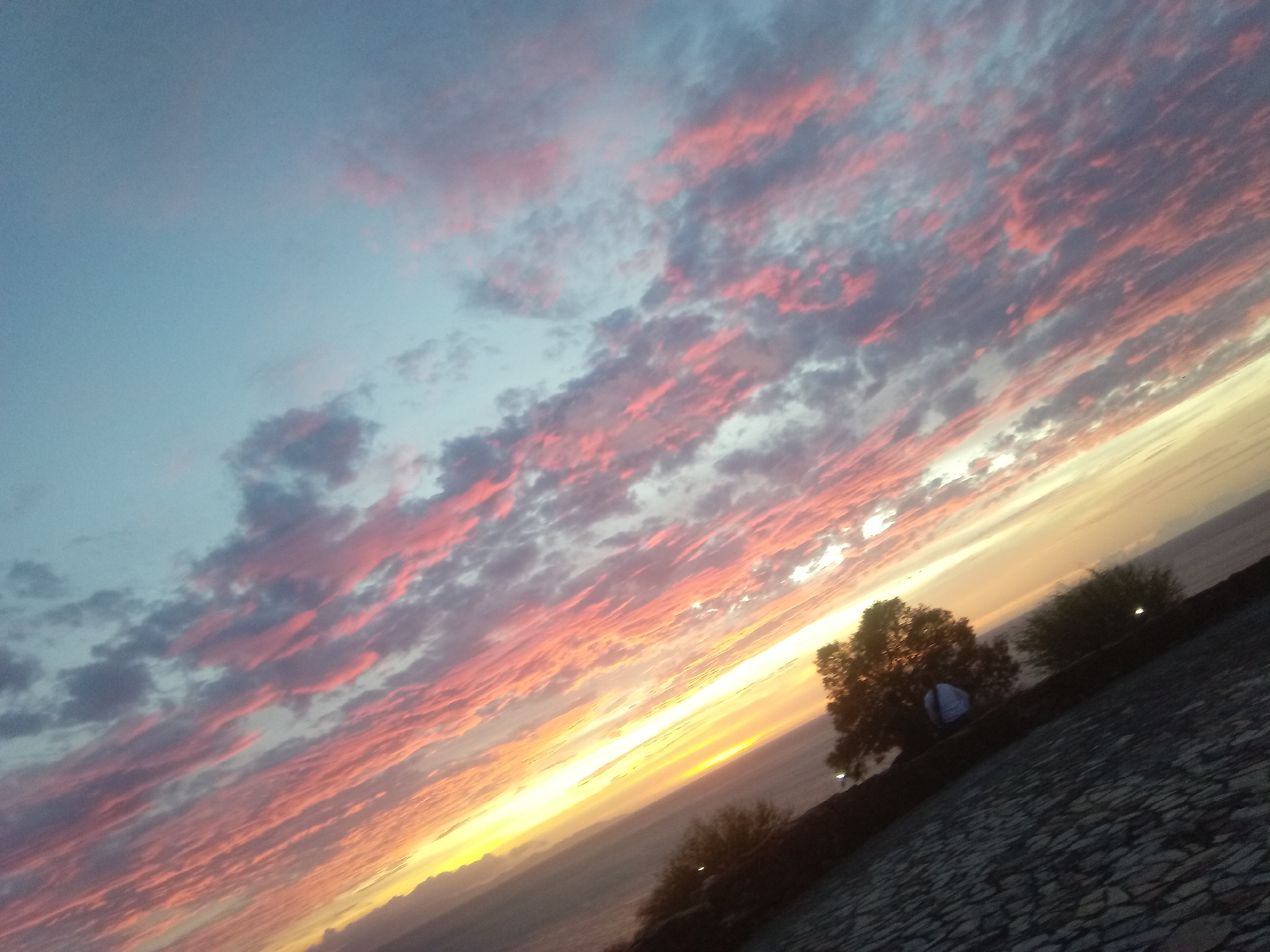 The photograph captures a sunset over Juan Griego, Venezuela. The sky is filled with pink and grey clouds, radiating from a bright yellow horizon. In the foreground, there's a stone surface, possibly a pathway or rooftop, with some vegetation including trees and bushes. A person in a light-colored shirt stands near the trees. In the distance, a body of water stretches to meet the sunset.