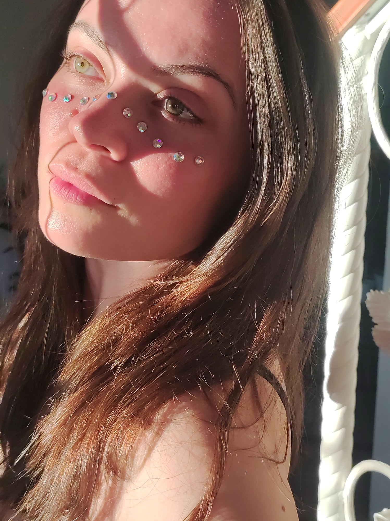 A close-up shot shows a woman with fair skin, light brown hair, and a distinctive arrangement of small, iridescent gems on her face. The gems are placed along her cheekbones and around her left eye. One eye is green and the other is brown. Her lips are full and slightly parted. A strong, direct light source illuminates the left side of her face, creating sharp shadows. A white, ornate metal structure is partially visible in the background, suggesting an interior setting near a window. The focus is primarily on the woman's face and the play of light and shadow, creating a vibrant and striking image. The location Фоки, Russia is not discernible from the image.