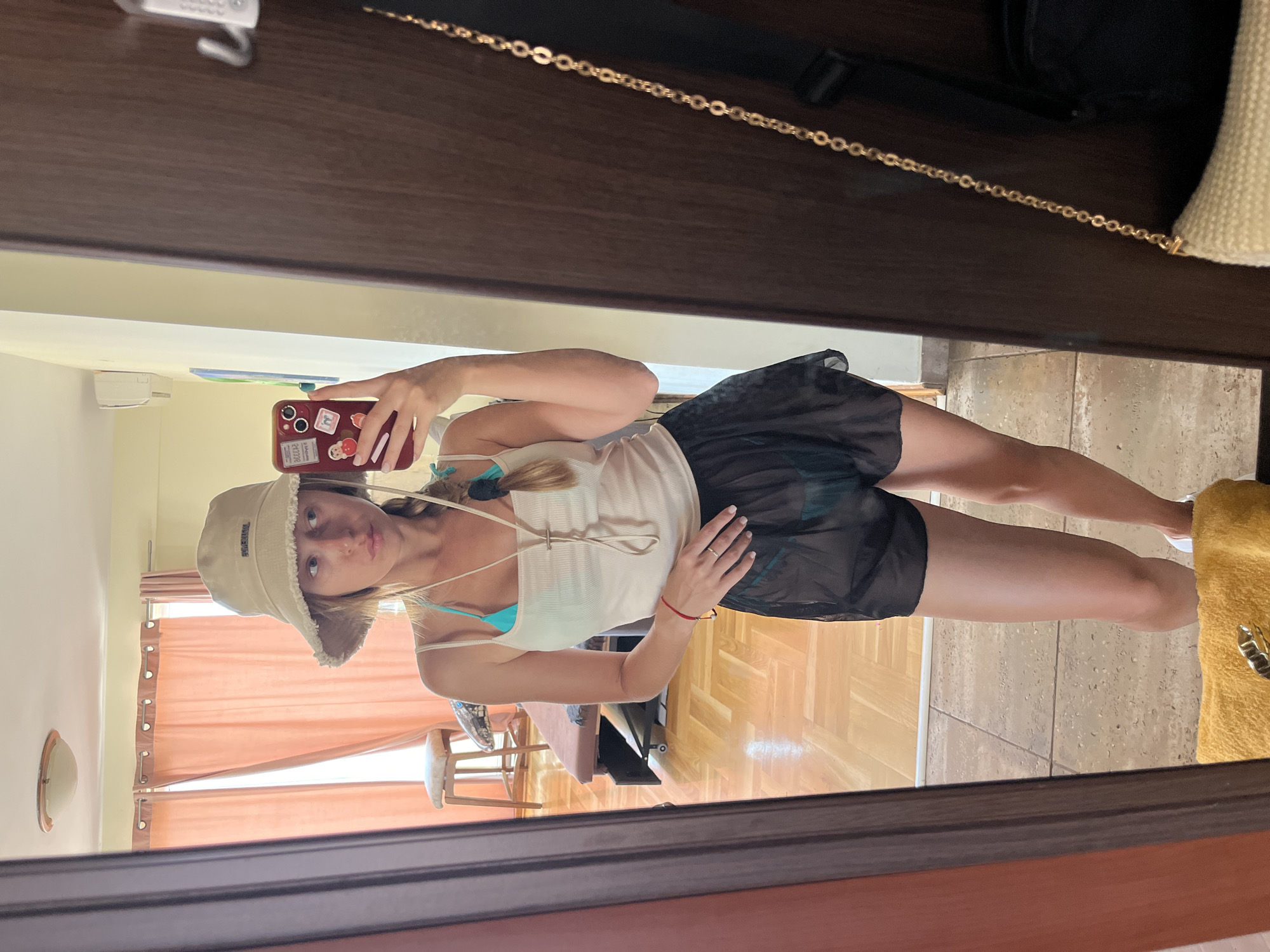 A young woman, viewed from the waist down, stands in a room and takes a mirror selfie. She is wearing a light-colored tank top over a turquoise bikini top, sheer black shorts, and white sneakers. A light-colored bucket hat with a frayed brim adorns her head. Her blonde hair is styled in a braid. A red cell phone with stickers is held in her right hand, pointed towards the mirror. Behind her, a wooden floor with a parquet pattern is visible, along with peach-colored curtains hanging in a window and a stool. A dark brown door frame surrounds the mirror. A gold chain purse strap hangs to the right of the woman.