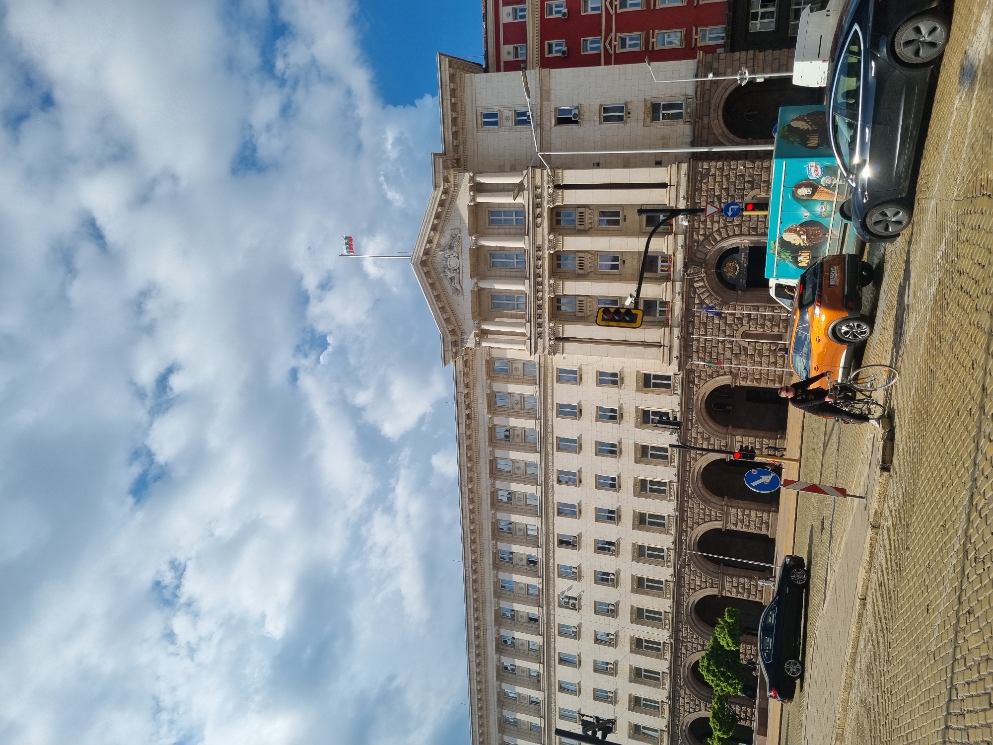 The image shows a street scene in Sofia, Bulgaria. The architecture is dominated by a large, light-colored, ornate building, likely a government building, featuring rows of windows, arched doorways, and a flag on top. A red building is visible next to it. The sky is partly cloudy, with blue sky peeking through. On the right side of the image, there is a cobbled street with parked vehicles, including a dark-colored car and an orange car. An ice cream truck is also present. A person is standing next to a bicycle. A traffic light is red. A street sign is visible.
