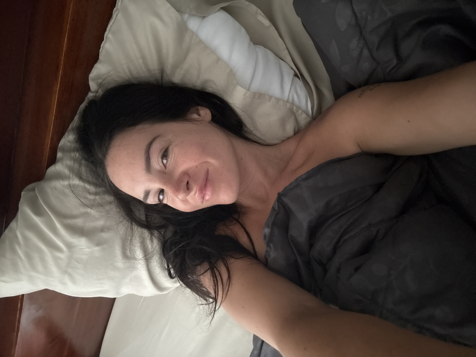 A woman with dark hair lies in bed, looking at the camera and smiling. She is covered by a dark grey blanket and resting her head on a light-colored pillow. The background shows a wooden headboard. The scene appears to be indoors, possibly in a bedroom, with natural light illuminating the woman's face. There are no discernible elements in the image that would suggest a location in North Kuta, Indonesia.
