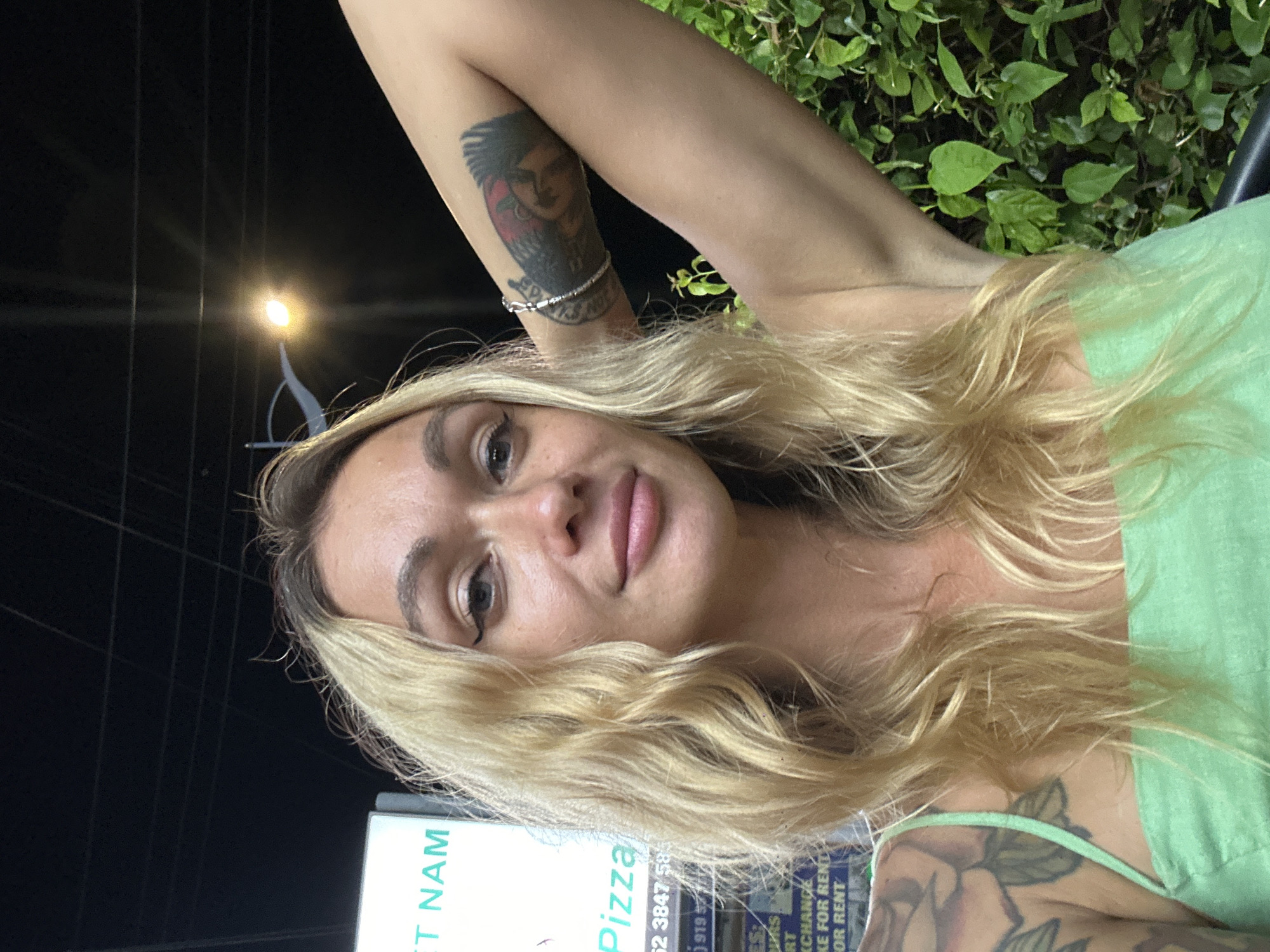 A woman with blonde hair is taking a selfie outdoors at night. She is wearing a light green top. She has tattoos on her arm and chest, including a portrait on her upper arm and floral designs on her chest. In the background, there are green bushes, a street lamp, and a sign that reads "Pizza" and "FOR RENT." The sign also includes a phone number. The overall impression is casual and relaxed.