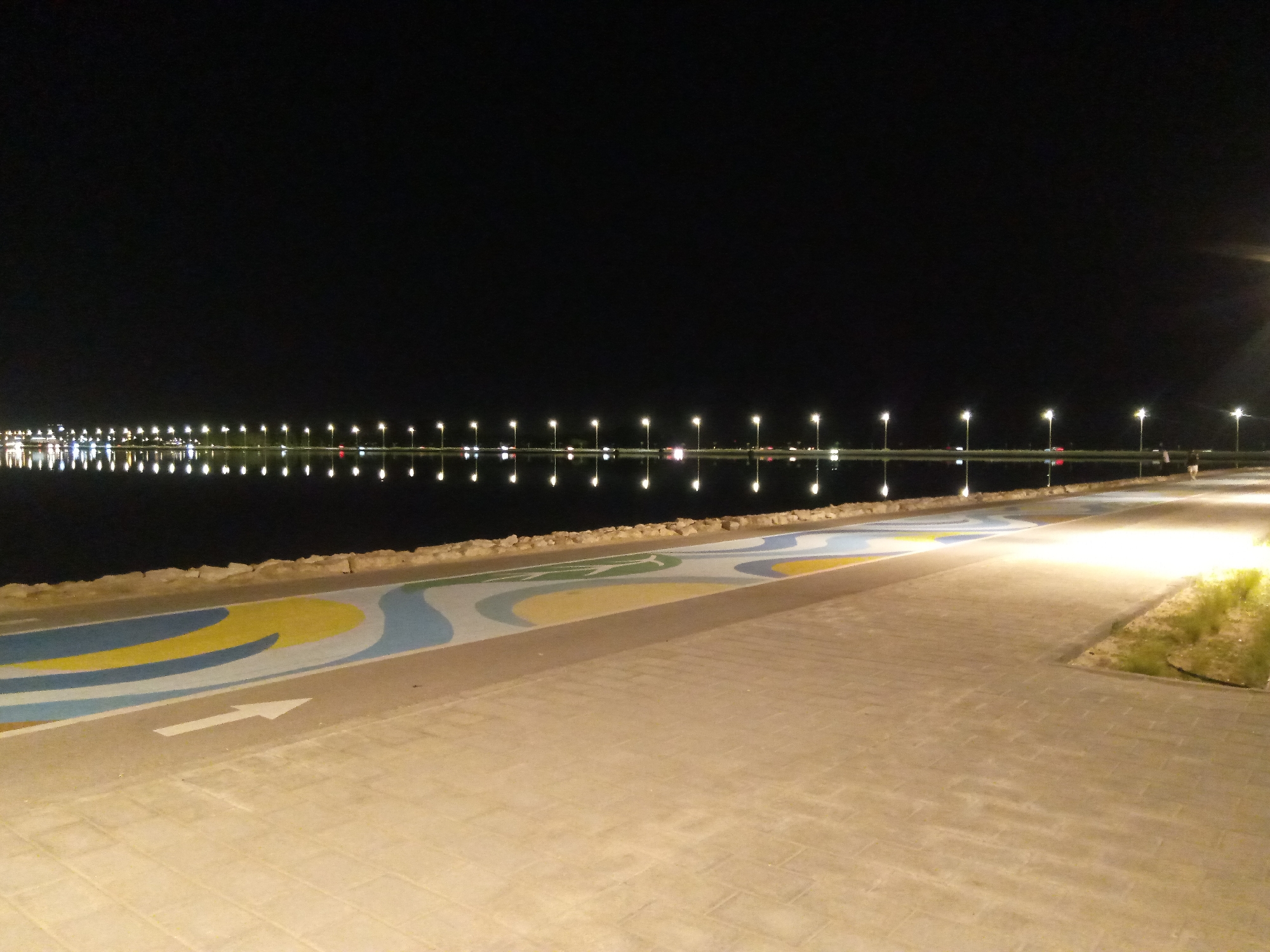 This image captures a nighttime scene along the waterfront in Al Khobar, Saudi Arabia, likely at Al Khobar Corniche or a similar coastal area. The foreground features a paved walkway with a decorative, wavy design in blue, yellow, and white. A white arrow indicating direction is visible on the walkway. Beyond the walkway, a low, rocky wall separates it from a body of calm water.
The water reflects a long, continuous line of streetlights that illuminate the distant shoreline. These lights appear as bright, distinct orbs, with their reflections creating a shimmering effect on the water's surface. The far background is dominated by darkness, with a few more distant lights suggesting the presence of buildings or a city skyline. The overall atmosphere is serene and modern, highlighting the well-lit urban landscape against the dark night sky. There are no identifiable people or specific objects in the immediate foreground or middle ground that are clearly discernible in detail.