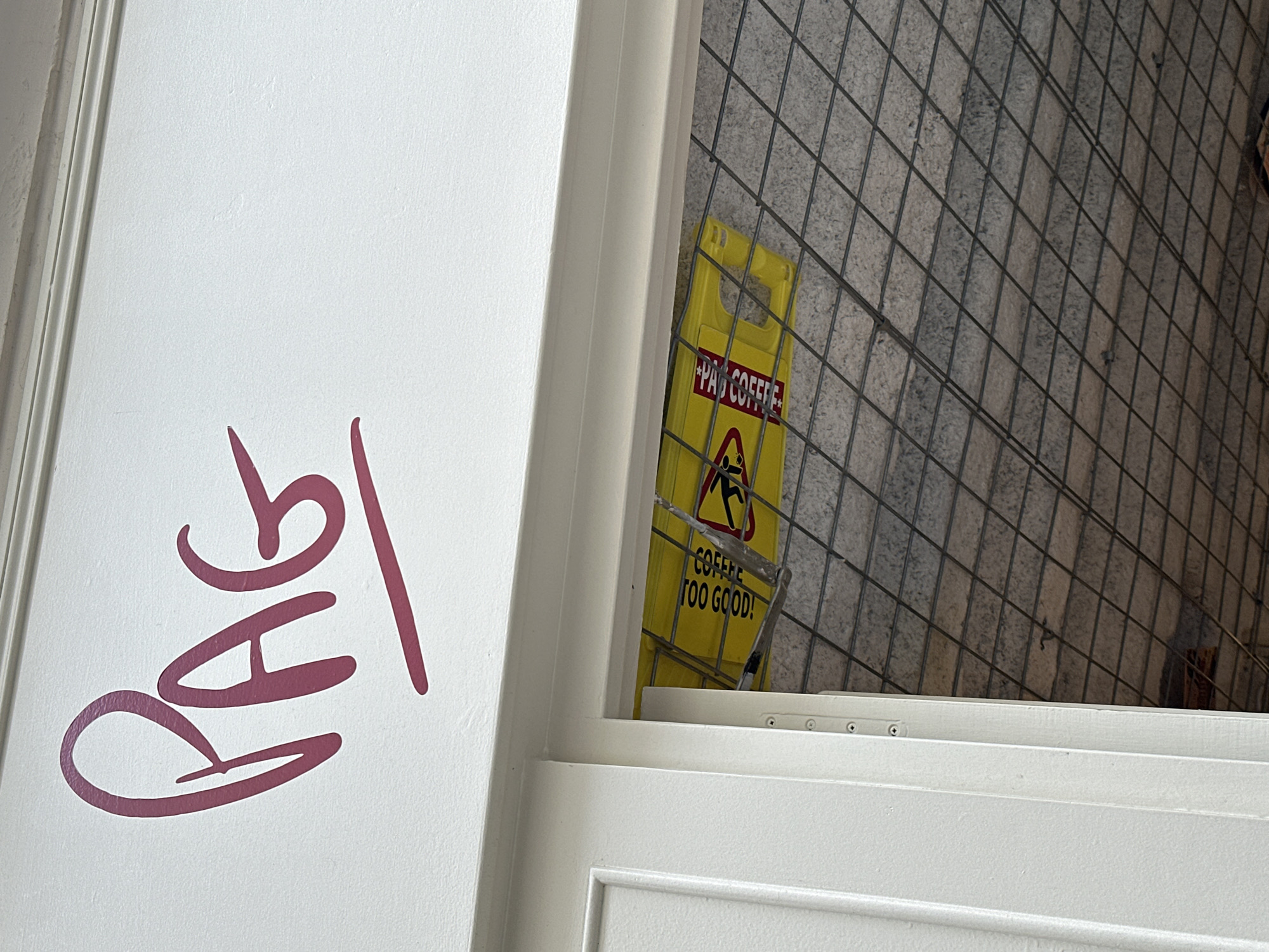 The image shows a white door partially open, revealing a "Wet Floor" sign on a stone-tiled surface. The sign is yellow and has the text "P*ss COFFEE" and "COFFEE TOO GOOD!". A red, handwritten inscription "BAG" is visible on the door. The location is Paris, France.