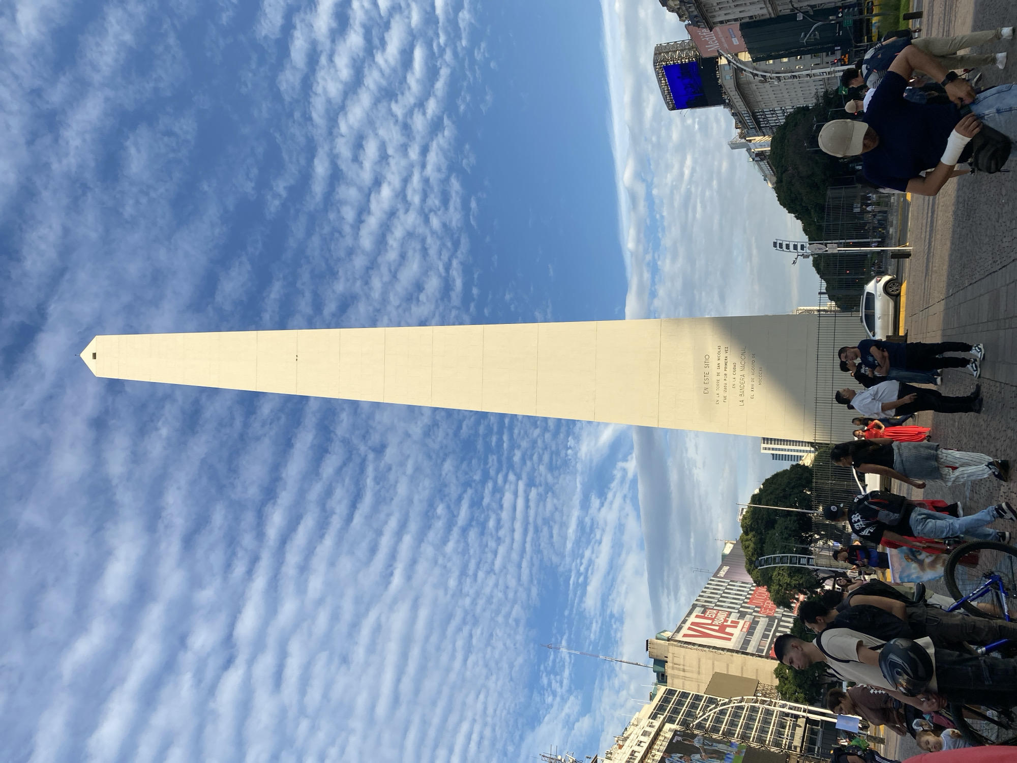 The image shows a tall, light-colored obelisk under a partly cloudy sky. The obelisk is the Obelisco de Buenos Aires, a historical monument in the city. The obelisk is surrounded by people and buildings. There are people standing and walking around the base of the obelisk. There is a street with cars, buildings, and trees visible. The sky is blue with white clouds.
