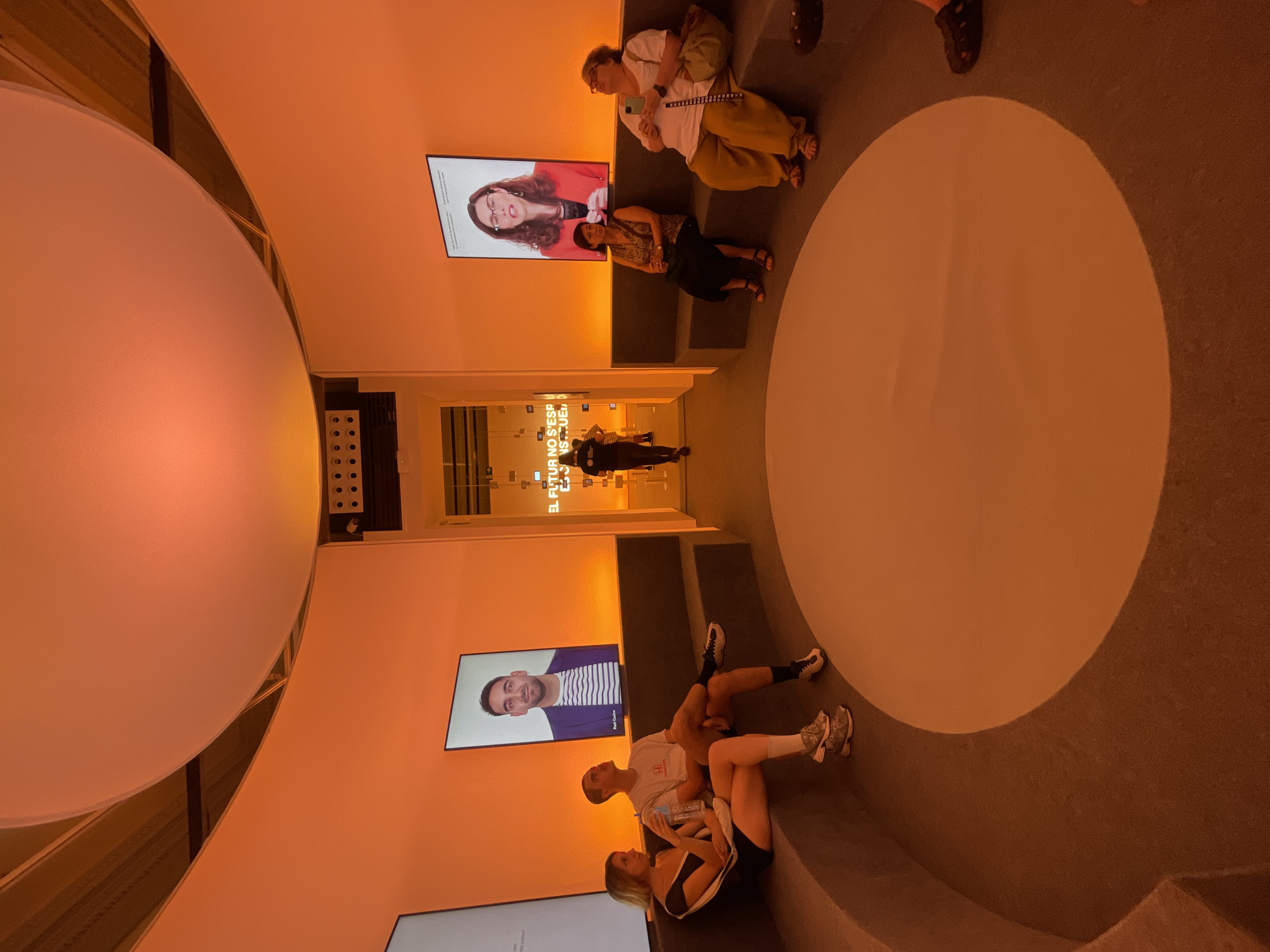 In Barcelona, Spain, within an exhibition space, a group of people are seated on a curved, built-in bench illuminated by warm orange light. The circular room features large screens displaying portraits and videos. On the left, a young couple sits together, with the woman in a black top and shorts, and the man in a white t-shirt and shorts. Behind them, a screen shows a man in a suit and striped shirt. To the right, two women are seated. One wears a patterned top and dark skirt, while the other is in a white t-shirt and tan pants. A screen above them shows a woman speaking, wearing glasses and a red blazer. In the background, a walkway leads to another area with text that appears to read "EL FUTURO NO S'ESCAPA" and a person is walking away. The floor is a dark carpet with a large, circular illuminated area in the center.