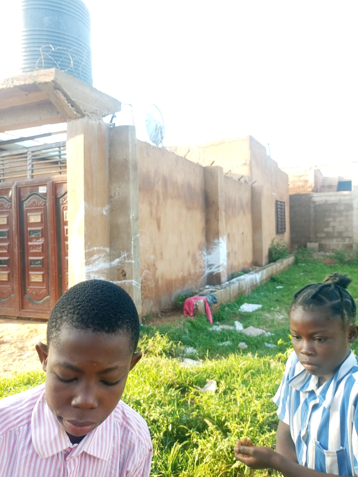 In Tempure, Nigeria, an outdoor scene captures two young children in the foreground. The child on the left, a boy, is wearing a pink and white striped collared shirt and is looking down with his eyes closed. To the right, a girl in a blue and white striped collared shirt with a pocket is looking towards the right with her hand outstretched. Behind them, a tan-colored wall dominates the mid-ground, with a drain or channel running along its base. There are scattered items of clothing, including a pink garment, on the ground near the wall. In the background, a large cylindrical water tank is visible, supported by a concrete structure. A metal gate with intricate designs is also present to the left of the wall. The environment appears to be a yard with green grass and some vegetation. The sky is bright and overcast.