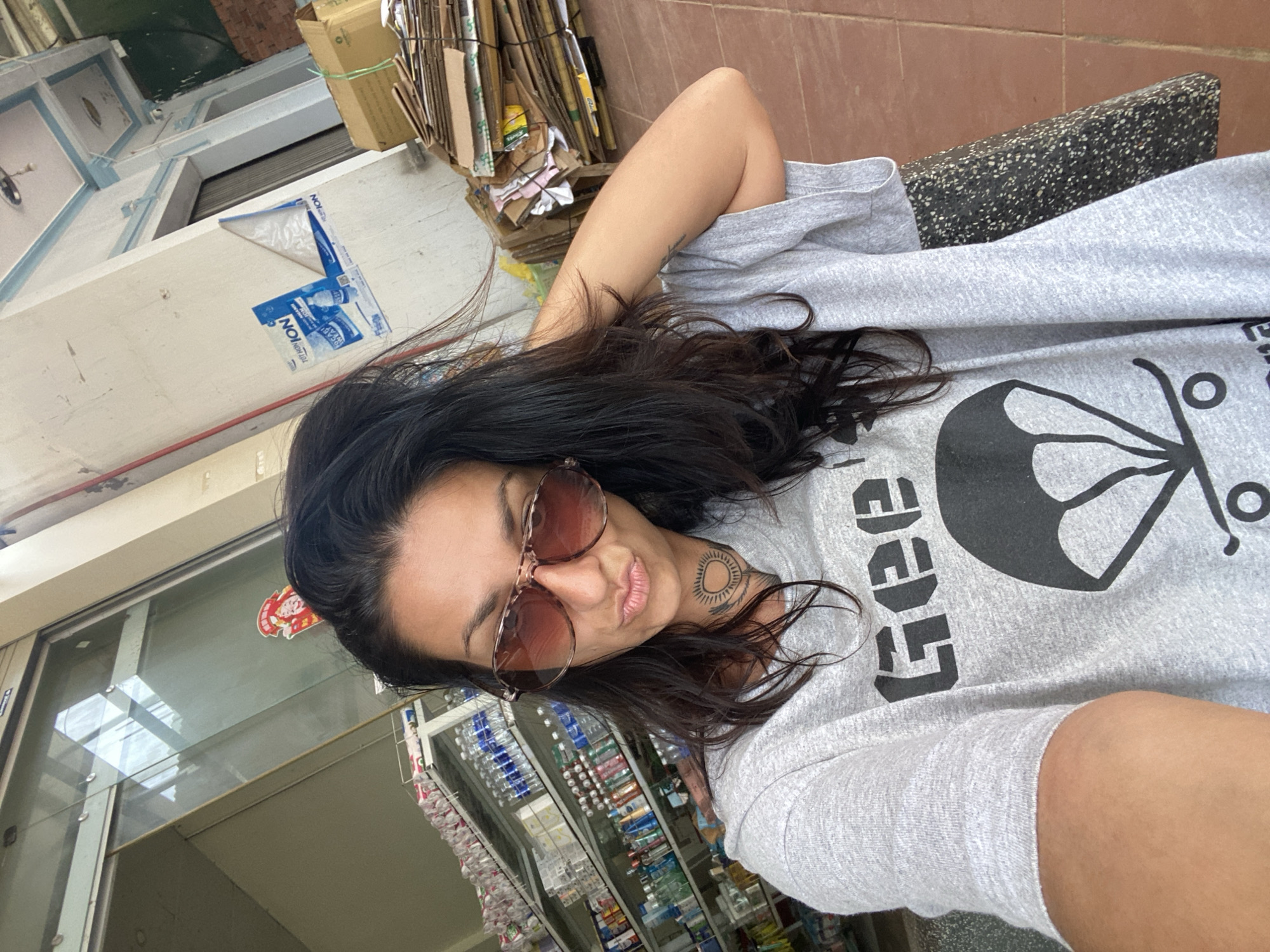 A woman with long, dark hair and sunglasses is taking a selfie in what appears to be a small shop or convenience store. She is wearing a gray t-shirt with a graphic print that includes the word "GEE" and a stylized parachute or umbrella over a skateboard. The background shows shelves stocked with various bottles and products, a roll-up metal door, a wall with posters, and a stack of cardboard boxes. The floor appears to be tiled. The location is identified as Thôn 2, Vietnam.