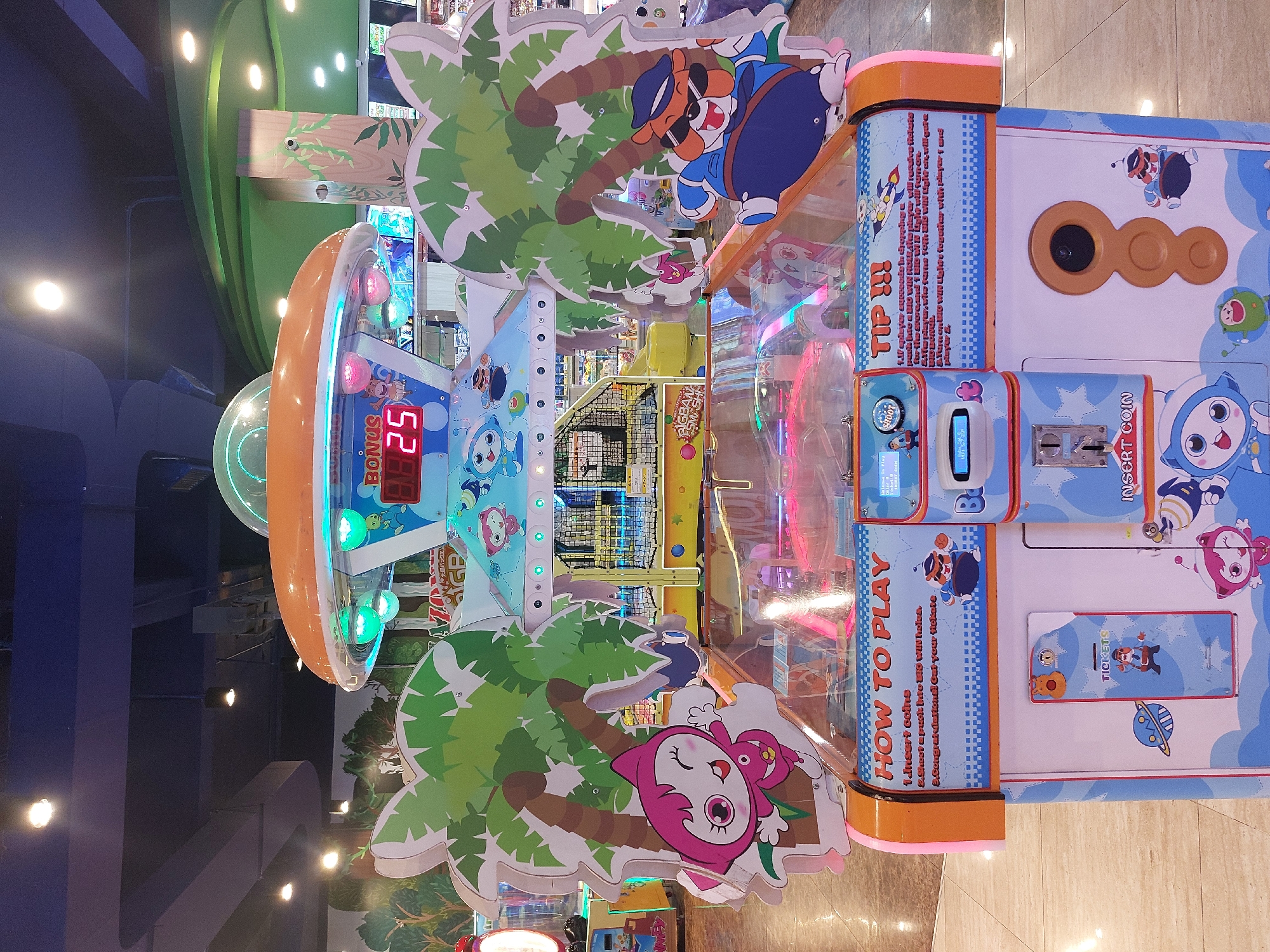 The image shows an arcade game machine, likely located in an entertainment center or mall in the City of Pematang Siantar, Indonesia. The machine is brightly colored, featuring cartoon characters like cats and a bear in a police uniform, as well as palm trees and a flying saucer design. The game appears to involve shooting pucks into holes. Instructions for playing, including inserting coins and collecting tickets, are displayed on the machine. The background suggests an indoor environment with ceiling lights and other arcade attractions visible.