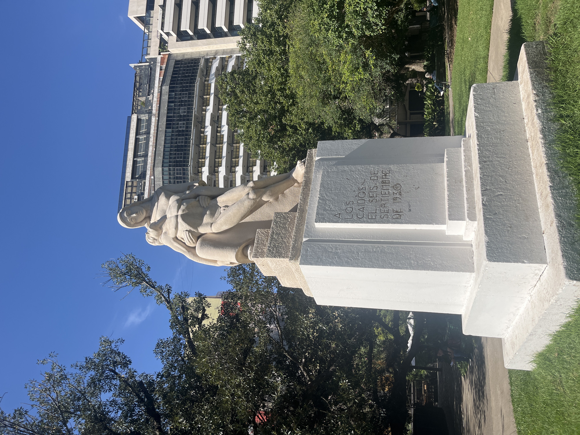 A large stone monument stands prominently in what appears to be a city park under a clear, bright blue sky. The monument consists of a multi-tiered, stark white base with an allegorical sculpture group mounted on top. The sculpture, crafted from light-colored stone, depicts several intertwined human figures, suggesting struggle, support, or a commemorative embrace.
Inscribed on the front face of the white pedestal is Spanish text that reads: "A LOS CAIDOS EL SEIS DE SEPTIEMBRE DE 1930" (To the Fallen on September Sixth, 1930). This inscription indicates the monument commemorates an event or individuals from that specific date.
Surrounding the base of the monument is neatly trimmed green grass, and lush green trees frame the scene, suggesting a park-like setting common in Buenos Aires, Argentina. In the background, a modern high-rise building with a light-colored facade and numerous dark windows and balconies rises towards the sky, contrasting with the classical monument. Another building is partially visible behind some trees. The bright lighting suggests it is daytime, likely mid-morning or afternoon, with strong sunlight casting clear shadows. A paved path is visible in the distance, and faint figures of people sitting on a bench can be discerned further back, reinforcing the public park environment.