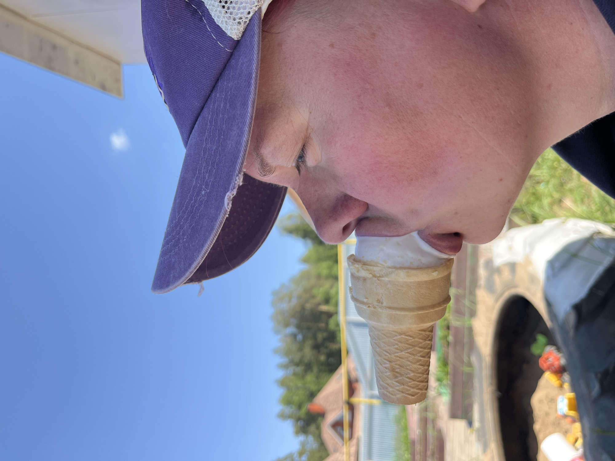 A person wearing a purple baseball cap is eating an ice cream cone. The scene is outdoors. The person's face is flushed. In the background, there is a blue sky and trees. There is also a building, possibly a house, and a tire that is likely a part of a playground. The exact location within Chaikovsky, Russia, cannot be determined without additional information.