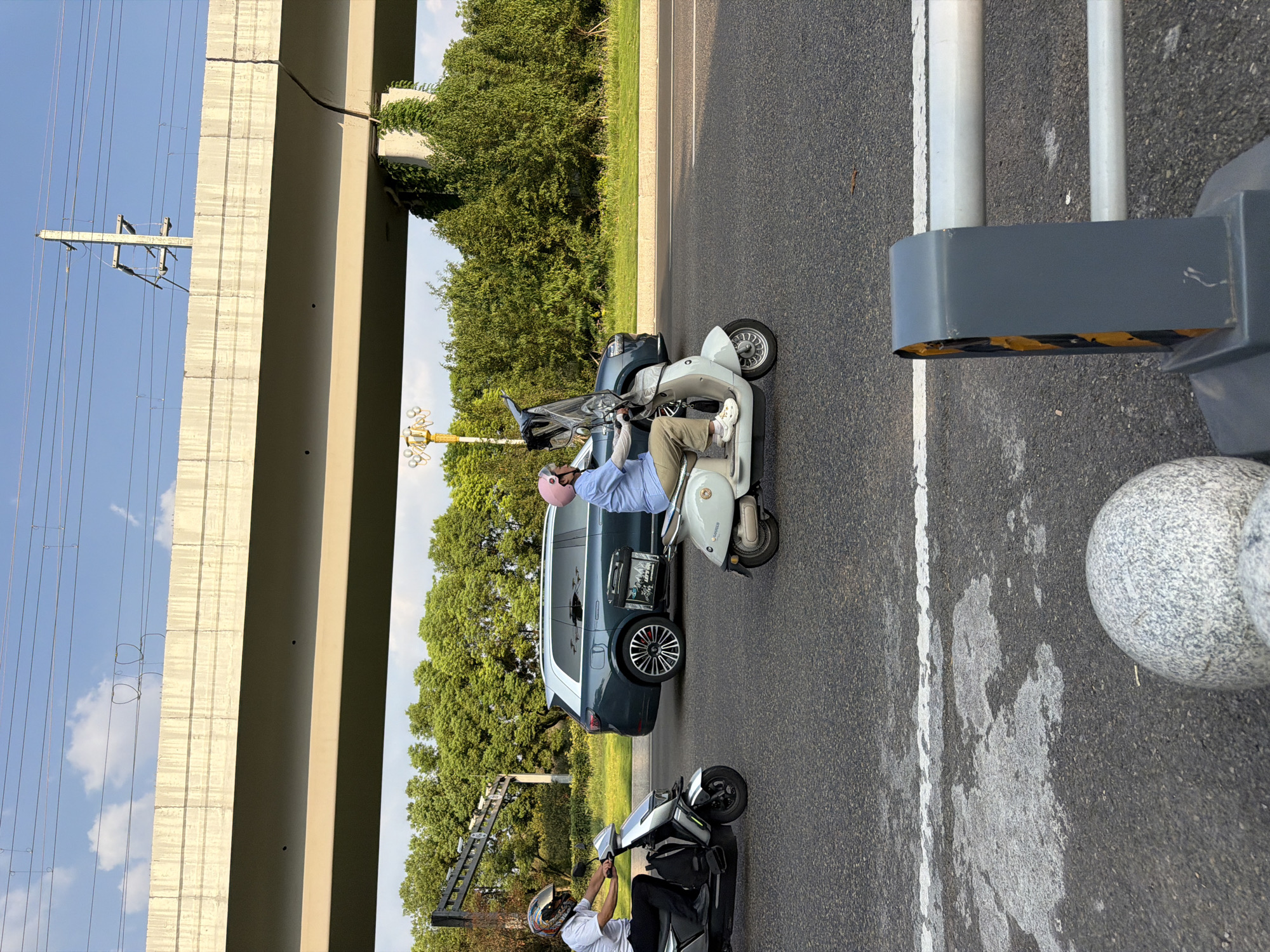 In the city of Jiangjin, China, on a sunny day with a blue sky and scattered clouds, two individuals are seen riding electric scooters on a paved road. The road has clear white lane markings and some wet patches. In the background, a multi-lane overpass or bridge dominates the upper portion of the image, with power lines strung above it. Below the overpass, a line of lush green trees forms a natural barrier.
On the left side of the road, a person wearing a white shirt and a helmet is riding a silver electric scooter. They are looking forward, navigating the road. To the right of the first scooter, a second individual, also wearing a helmet (pink) and a light blue shirt, is riding a white electric scooter. This scooter has a transparent windshield. The person appears to be looking ahead, focused on their ride.
Behind the scooters, a dark blue SUV with tinted windows is driving in the same direction. The SUV has distinctive silver rims and a sunroof.
In the foreground, partially visible, are elements of street infrastructure. On the right, a grey and yellow traffic barrier is present, featuring reflective elements. On the left and center foreground, there are two large, grey, spherical objects, possibly decorative bollards or planters. The overall scene depicts a common urban commute on a clear day.