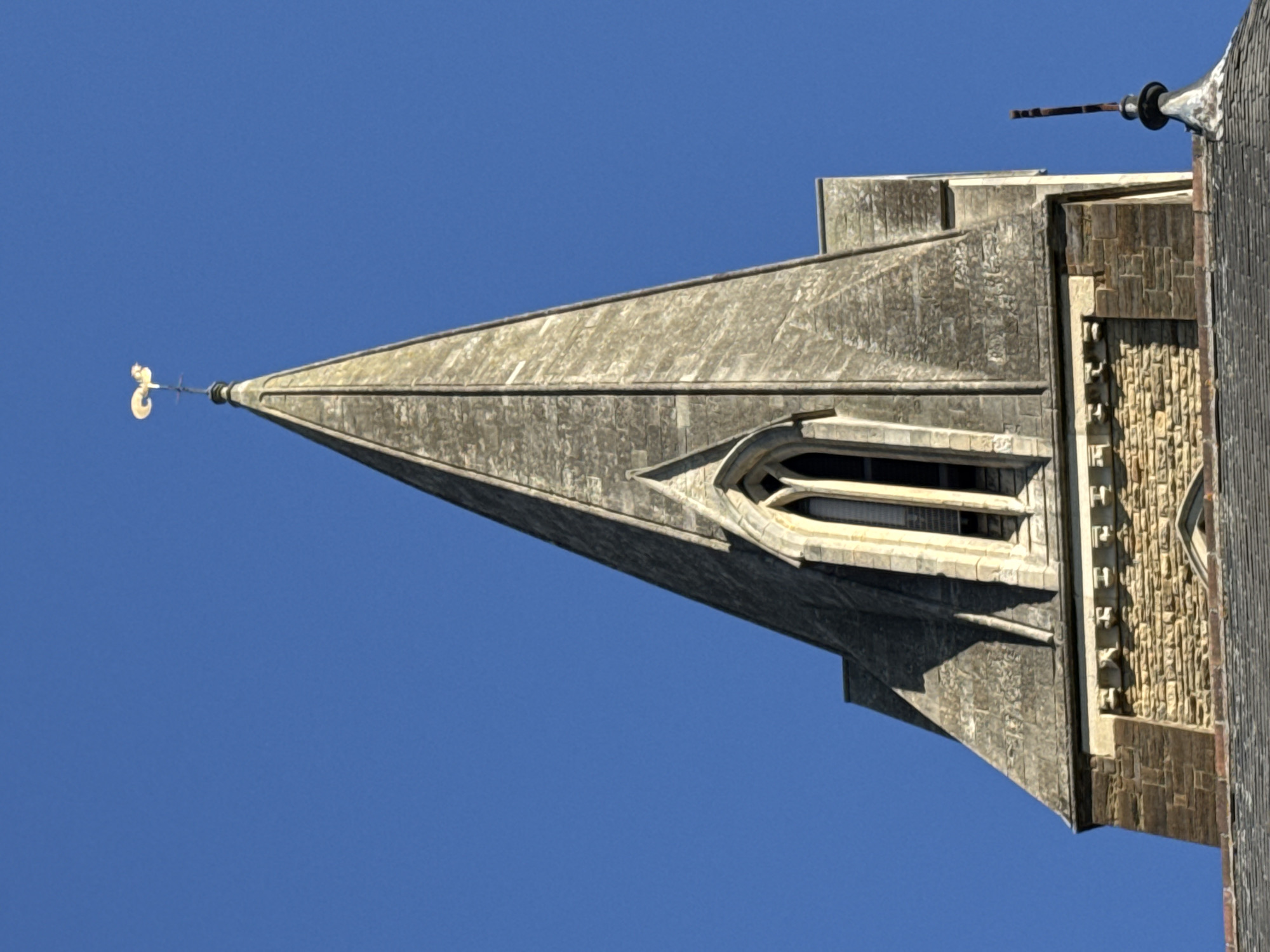 The image features a detailed, low-angle view of a stone spire or steeple against a bright, clear blue sky. The spire is constructed from textured, weathered grey and beige stone blocks, displaying architectural features typical of older, possibly ecclesiastical buildings.
At the very top of the spire, a decorative, golden-colored weather vane is mounted, appearing as a stylized '2' or 'S' shape with a small arrow. Below this, approximately halfway down the visible portion of the spire, a tall, arched window is set into the stone. This window features intricate gothic or neo-gothic detailing in lighter-colored stone, framing dark interior glass or louvers.
To the right of the spire, a section of the main building is visible, showcasing additional textured stone work. This section consists of various patterns of rectangular and unevenly cut stone blocks, contributing to a rugged, historic aesthetic. A dark roofline is also discernible on the right side.
The sky is a uniform, deep blue, devoid of clouds, indicating bright, sunny weather. The strong sunlight creates clear highlights and shadows on the stone work, accentuating its texture and depth. The photograph is taken looking upwards at the structure and is displayed in a sideways orientation, making the spire appear to point horizontally to the left. No human activity or visible text is present in the image. Given the location context of Far Cotton, United Kingdom, the architecture likely represents British style.