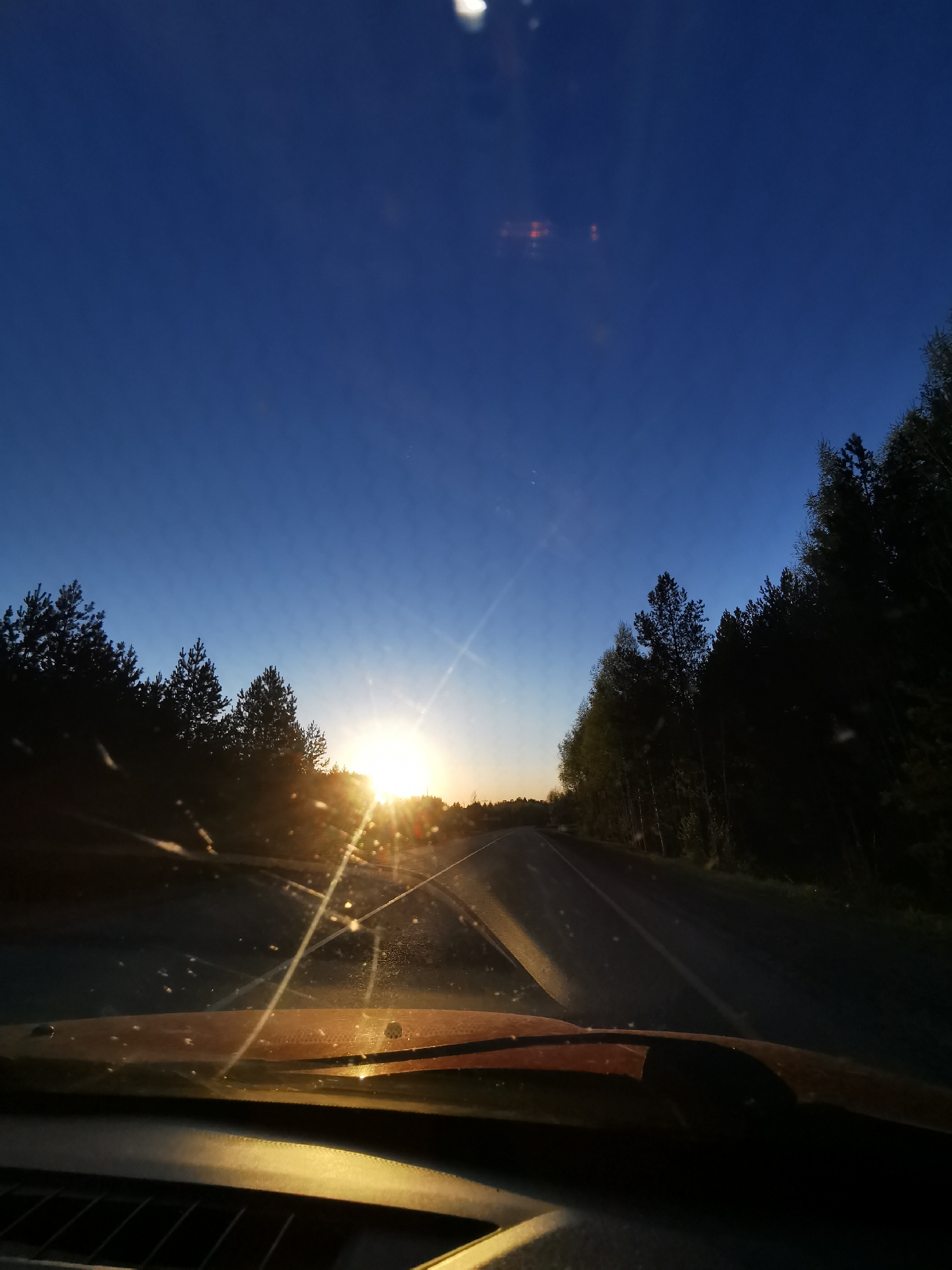 A forward-facing view from inside a vehicle shows a paved road leading into a sunrise or sunset. The road is flanked by trees on either side, silhouetted against the colorful sky. The interior of the vehicle, including the dashboard and part of the windshield with a wiper, is visible in the foreground. The image was taken in the vicinity of the city of Chaikovsky, Russia.