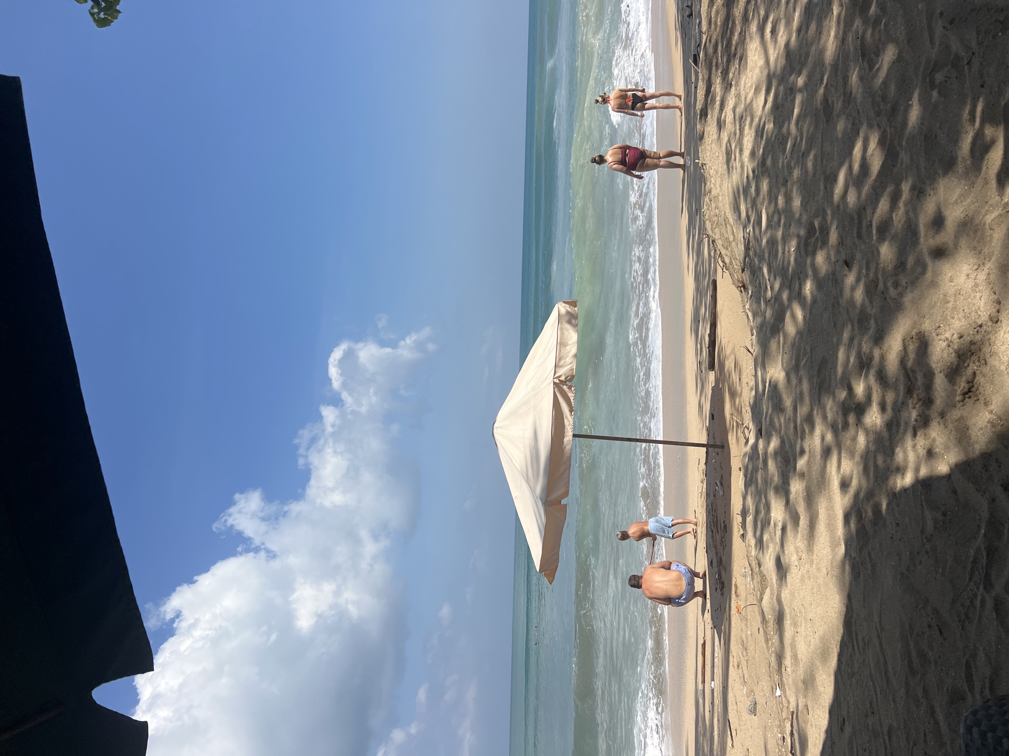 A sunny day at the beach in Kuta, Indonesia, features a sandy shore with gentle waves lapping the coast. The turquoise ocean stretches towards the horizon under a bright blue sky dotted with white clouds. In the foreground, a large, cream-colored beach umbrella provides shade.
Two individuals, a man in blue shorts and a child in blue shorts, are near the water's edge. The man is crouching, and the child is standing, holding a long stick. Further down the beach, two women in bikinis face the ocean. One woman wears a burgundy bikini, and the other wears a black bikini.
The sand is scattered with debris, including plastic bottles and driftwood. Shadows from unseen foliage cast dappled patterns across the sand. The scene conveys a relaxed atmosphere of a typical beach day in Kuta.