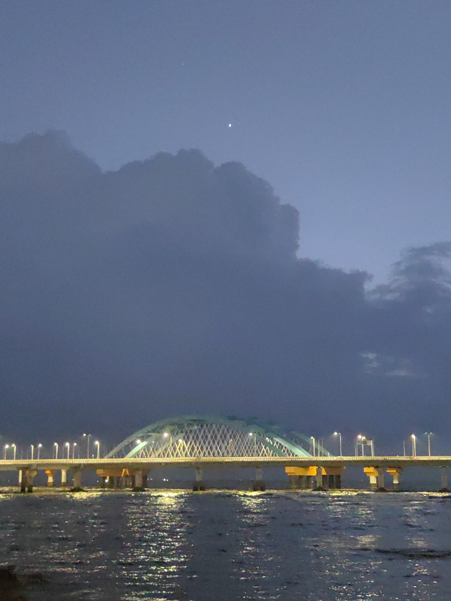 A bridge with arching supports and diamond-shaped netting spans across dark water, illuminated by rows of streetlights and reflected lights on the water's surface. The sky above is a deep twilight blue, partially obscured by large, dark clouds. A single bright star or planet is visible high in the sky. The scene appears to be a coastal area, possibly in Mumbai, India, given the architectural style of the bridge and the overall atmospheric conditions.