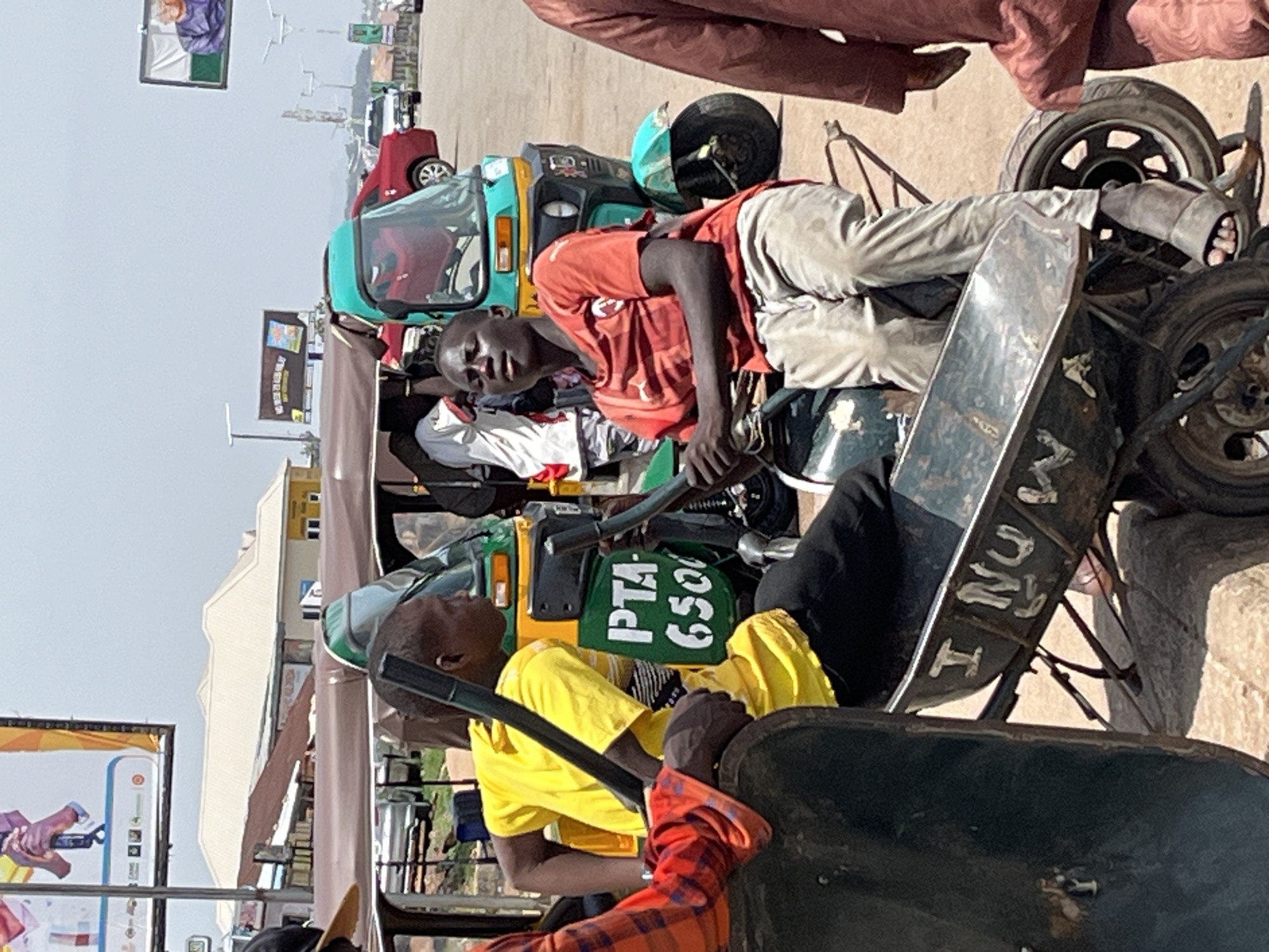 An outdoor scene in Gura Top, Nigeria, depicts a busy street or market environment during what appears to be a sunny day, with clear skies overhead. The image is oriented vertically (rotated clockwise 90 degrees from standard viewing).
In the mid-ground, a young dark-skinned man wearing a red short-sleeved shirt and light-colored trousers is resting or lying across the back seat of a teal and yellow motorized tricycle (commonly known as a keke napep in Nigeria). He looks directly towards the camera with a neutral expression. Behind him, another similar tricycle is visible.
Further into the foreground, another dark-skinned man in a bright yellow short-sleeved shirt is seated in the driver's position of a tricycle, looking away from the camera. On the back of his tricycle, a green fabric covering displays white text: "PTA 6500". A portion of a person in a red and black plaid shirt is visible in the extreme bottom right corner. Another person's legs, clad in reddish-brown fabric, are visible at the top edge of the frame, possibly standing near or on another vehicle.
A black metal structure, likely part of a vehicle or a cart, runs across the lower mid-ground, bearing white lettering that appears to read "I NU" followed by blurred characters such as "K9" or "K7". In the background, several other vehicles, including a red car, are parked or driving. Distant buildings are visible under the bright sky.
Two large billboards are present in the background. One billboard, to the left, features an older person in a purple garment alongside what appears to be the green and white vertical stripes of the Nigerian flag, possibly a political advertisement. Another billboard further to the right shows graphics of hands holding an object. The overall scene captures a moment of daily life and transportation in a Nigerian locality.