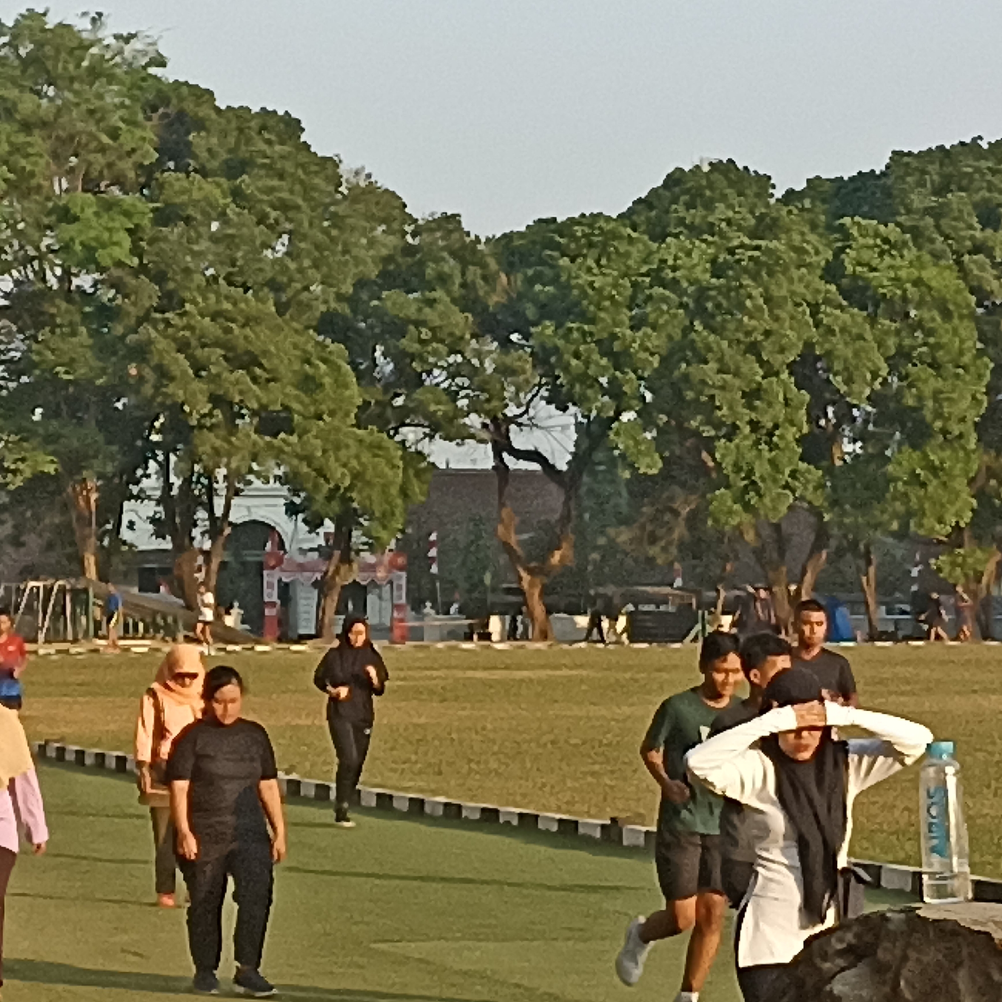 The image depicts an outdoor scene in Magelang, Indonesia, likely at Alun-Alun Kota Magelang, a popular public space. Several people are engaged in various activities on a grassy field, bordered by a black and white striped track. In the foreground, a woman in a white shirt and black hijab is covering her eyes with her hands, while two young men in athletic wear are jogging behind her. Further back, a woman in a black hijab is jogging on the track, and another woman in an orange hijab and a woman in a black shirt are walking. In the background, large, mature trees with dense green foliage dominate the scene, with a white, arched building with red accents visible behind them, possibly part of a historical structure or event venue. The lighting suggests it is either early morning or late afternoon.