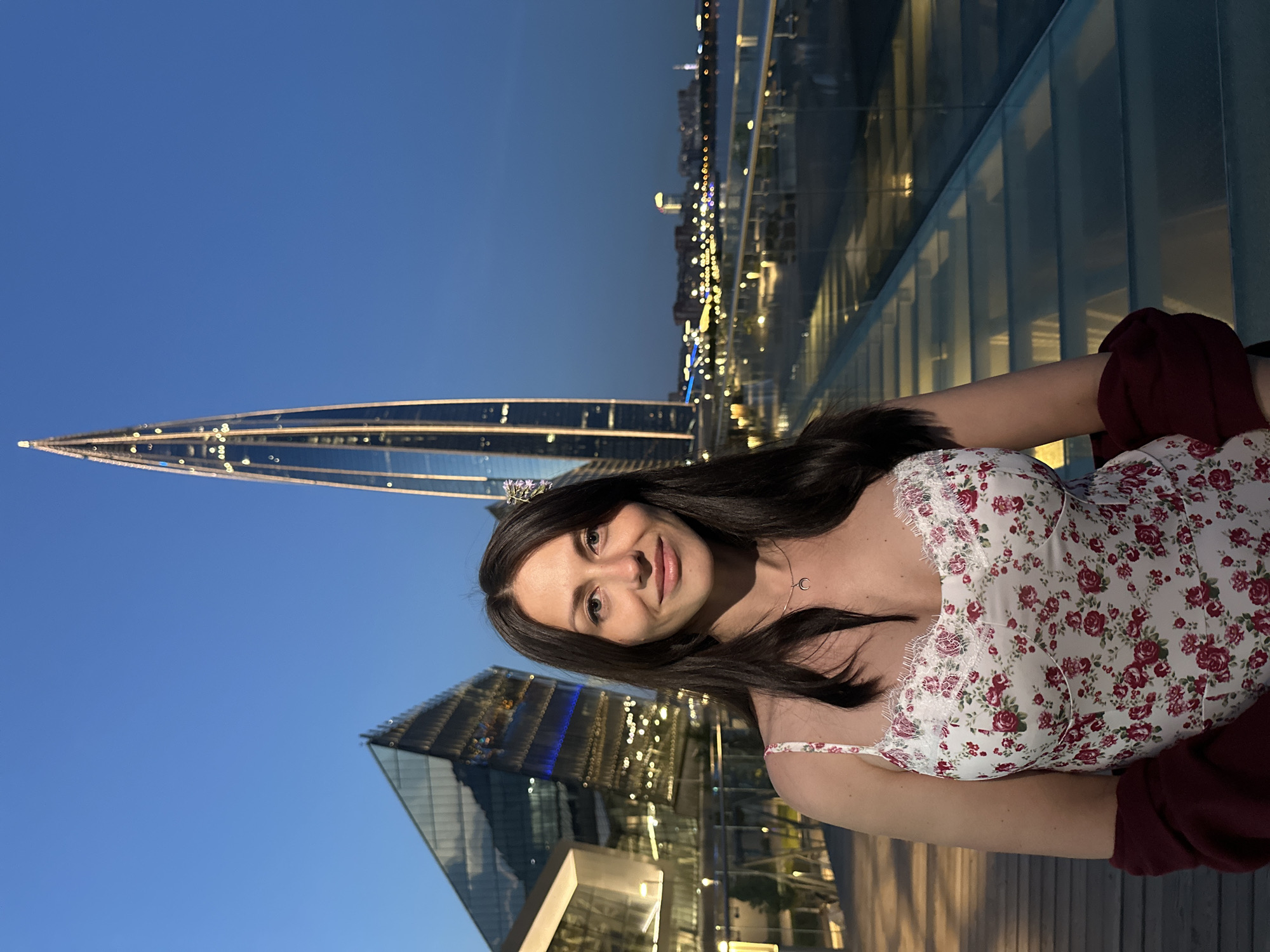 A woman with long, dark hair is posing for a photo in front of a modern cityscape at dusk. She is wearing a white floral dress with a lace trim and a crescent moon necklace. Behind her, a tall, pointed skyscraper, the Lakhta Center, dominates the skyline. To the left, a geometric glass building is visible. The scene appears to be on an elevated walkway or observation deck overlooking a body of water and a city skyline illuminated by lights. The sky is a deep blue, indicating the transition from day to night.