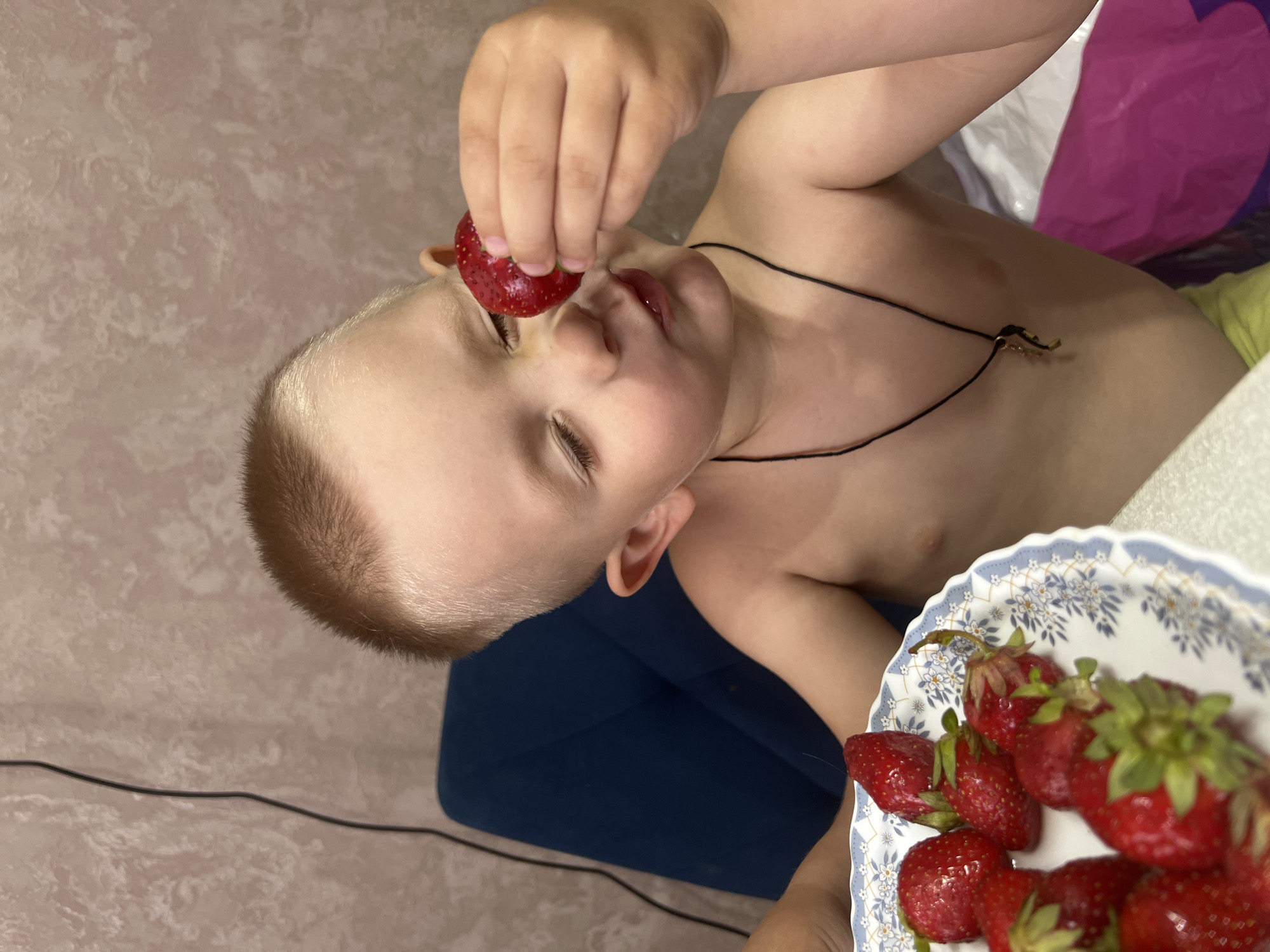 A young, shirtless boy with short, light brown hair is holding a strawberry up to his face with his right hand. He is looking at the strawberry with his eyes closed and his mouth slightly open. He is wearing a black cord necklace with a gold pendant. In front of him is a plate with several strawberries. The background consists of a textured, light pink wall and a dark blue chair. The scene appears to be indoors, possibly in a home environment. There is no identifiable information in the image to determine the specific location within Марковский, Russia.