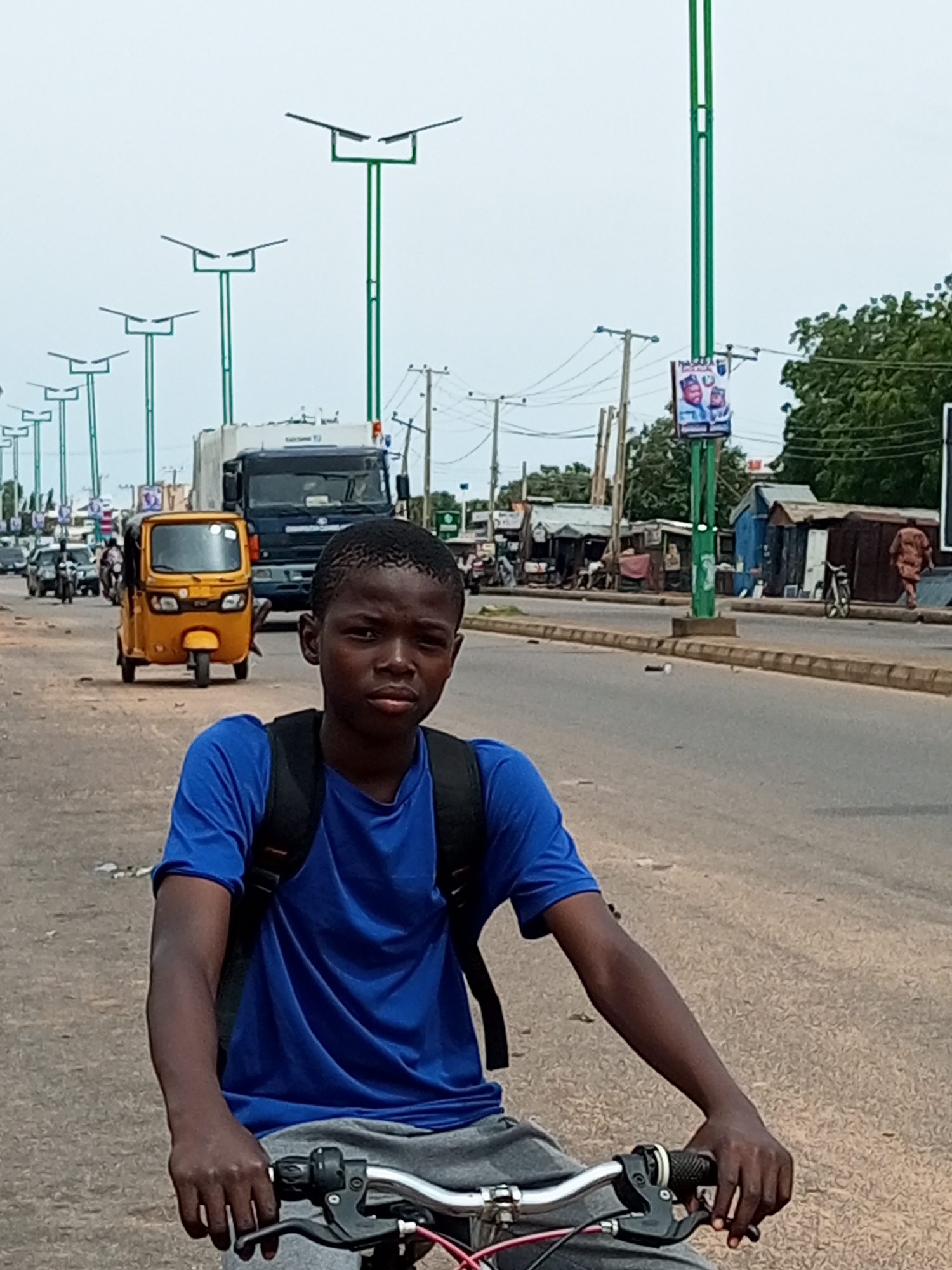 In Garko, Nigeria, a young boy wearing a blue t-shirt and grey shorts, with a black backpack, rides a bicycle on a dusty road. He is positioned in the foreground, facing the camera. Behind him, the road is populated with vehicles, including a yellow autorickshaw and a large truck. The scene depicts a busy street with various shops and stalls lining the sides. Several streetlights, some with solar panels, are visible along the road. The sky is overcast.