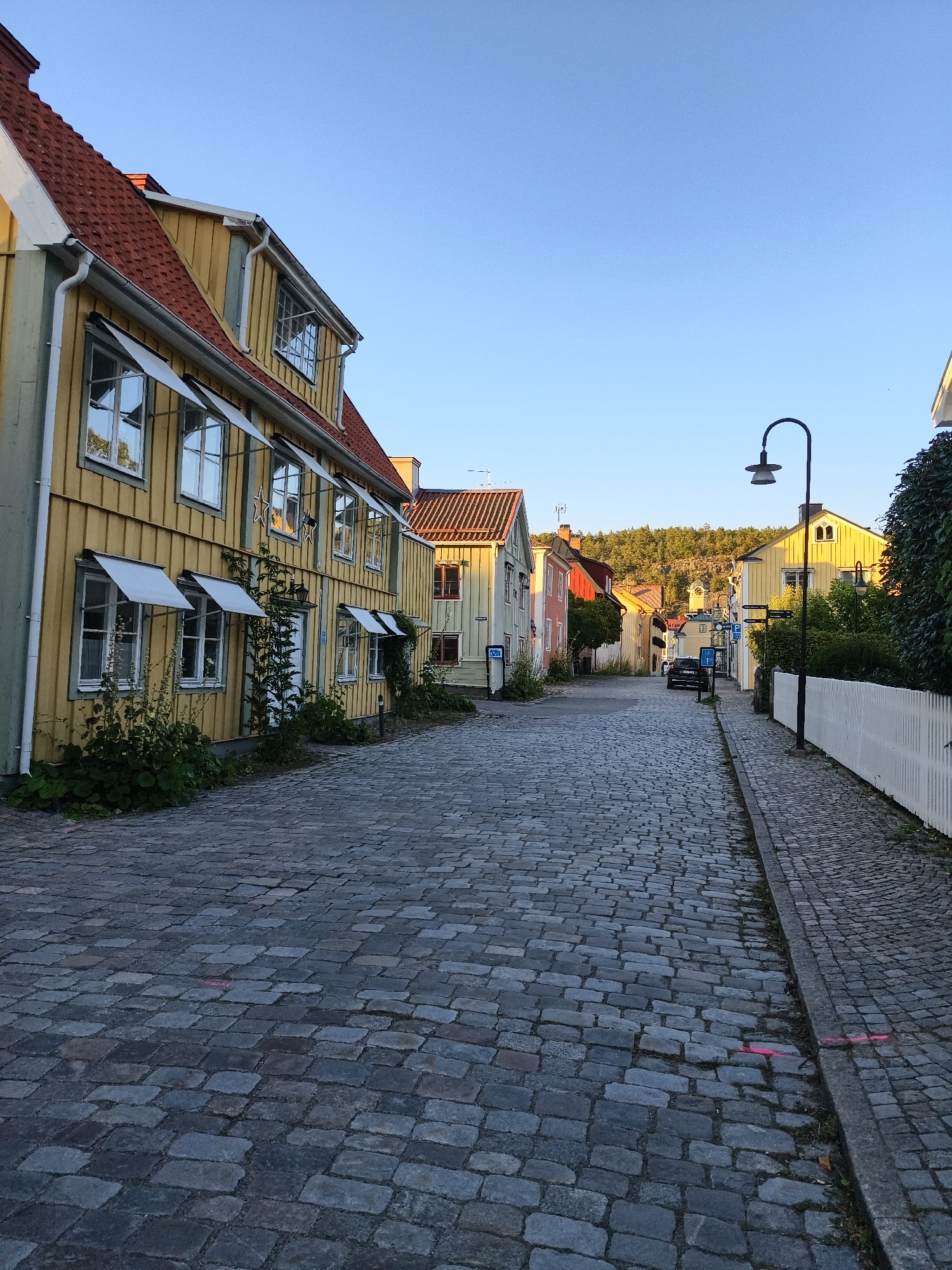 The image shows a view down a cobblestone street in Söderköping, Sweden. On the left side of the street are traditional Swedish wooden houses painted in yellow with red roofs. They have white-framed windows with awnings. Some houses have greenery growing up their sides. On the right side of the street, there is a white picket fence and a lamppost. Further down the street, more colorful houses are visible, and a car is parked. The street slopes gently upwards, leading towards a wooded hillside in the background under a clear blue sky. The lighting suggests it is either early morning or late afternoon. There are no people visible in the image.