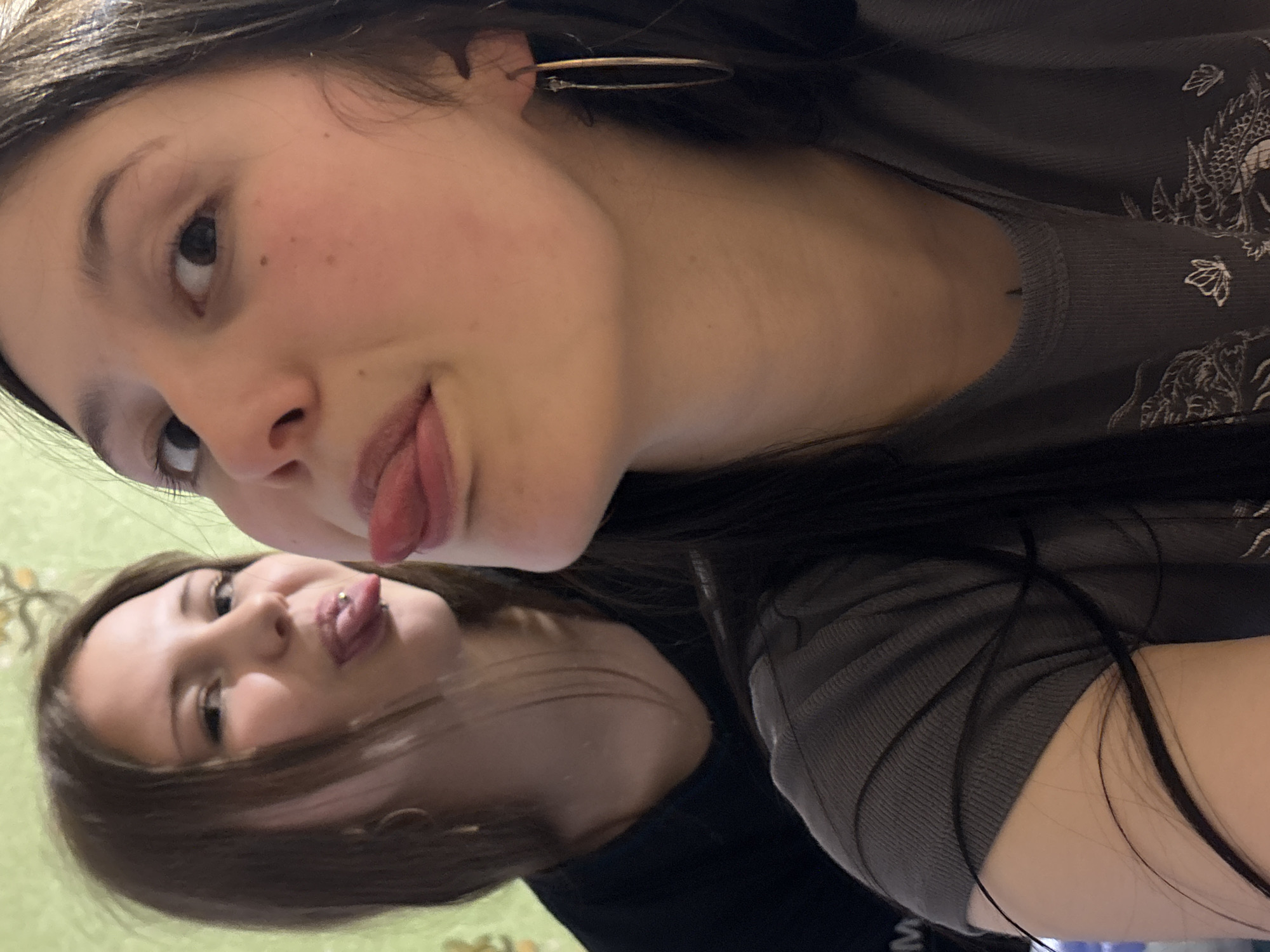 Two young women are positioned close together, faces angled toward the camera. The woman in the foreground has dark hair and is wearing a grey top with a dragon-like design. She has a gold hoop earring in her right ear and is sticking out her tongue. The woman behind her also has dark hair and is sticking out her tongue, which has a barbell piercing. They appear to be indoors, against a light green patterned wall. No information is provided in the image that can support or refute the supposed location within Фоки, Russia.