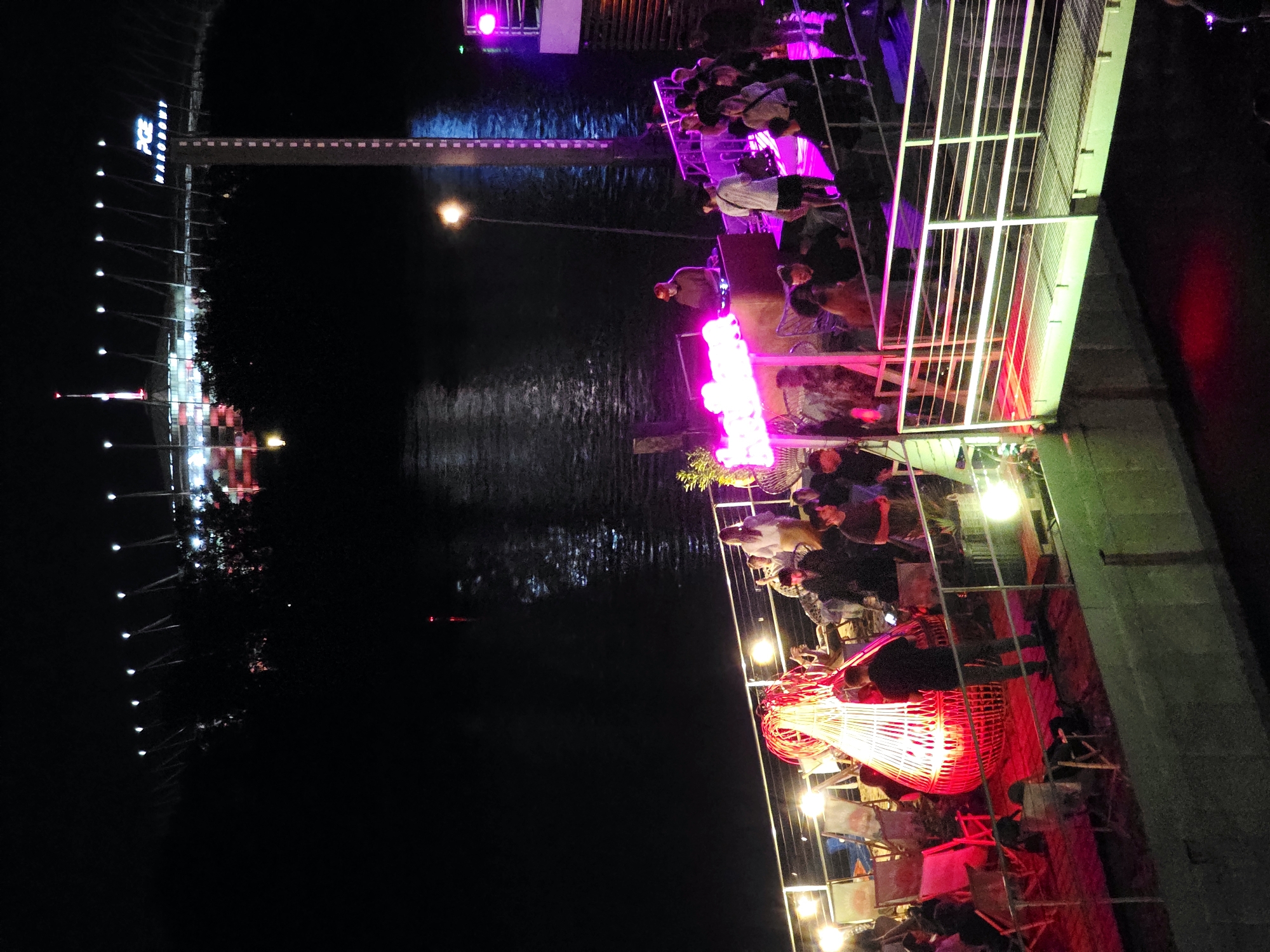 The image shows an outdoor nighttime scene near the Vistula River in Warsaw, Poland. In the background, the PGE Narodowy stadium is illuminated with bright lights. The foreground features a riverside venue with a deck area. Several people are visible on the deck, some standing and others seated. There is a DJ booth with a person operating it, and a crowd gathered around. Bright neon lights, primarily pink and red, illuminate the area. There are lounge chairs and a distinctive large, red, cone-shaped light fixture on the deck. The water of the Vistula River is visible, reflecting the lights and the dark night sky.