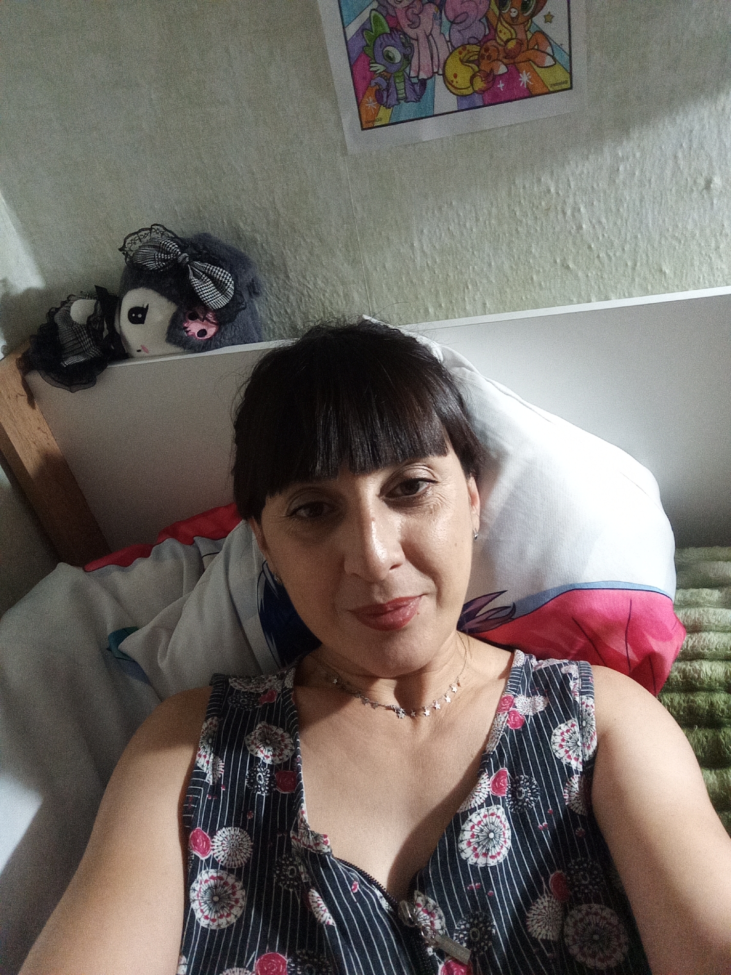 A woman is lying on a bed in what appears to be a bedroom. She has dark hair and is wearing a dark-colored dress with a floral pattern. There are pillows behind her, one of which has a pink and blue design. On a shelf above the bed, there are two stuffed animals, one gray with black and white accents and the other white with black and pink accents. A colorful drawing, possibly of cartoon characters, is hanging on the wall behind the stuffed animals. The walls appear to be painted a light green color and have a textured finish. The bedding is a mixture of white and green. The image is taken from a close-up perspective, focusing on the woman's face and upper body. The lighting suggests it might be daytime, possibly with natural light coming from a window. The overall environment appears to be domestic and casual. The provided location of Belogorsk, Russia, is noted, but there are no visual cues in the image that specifically confirm or deny this location.